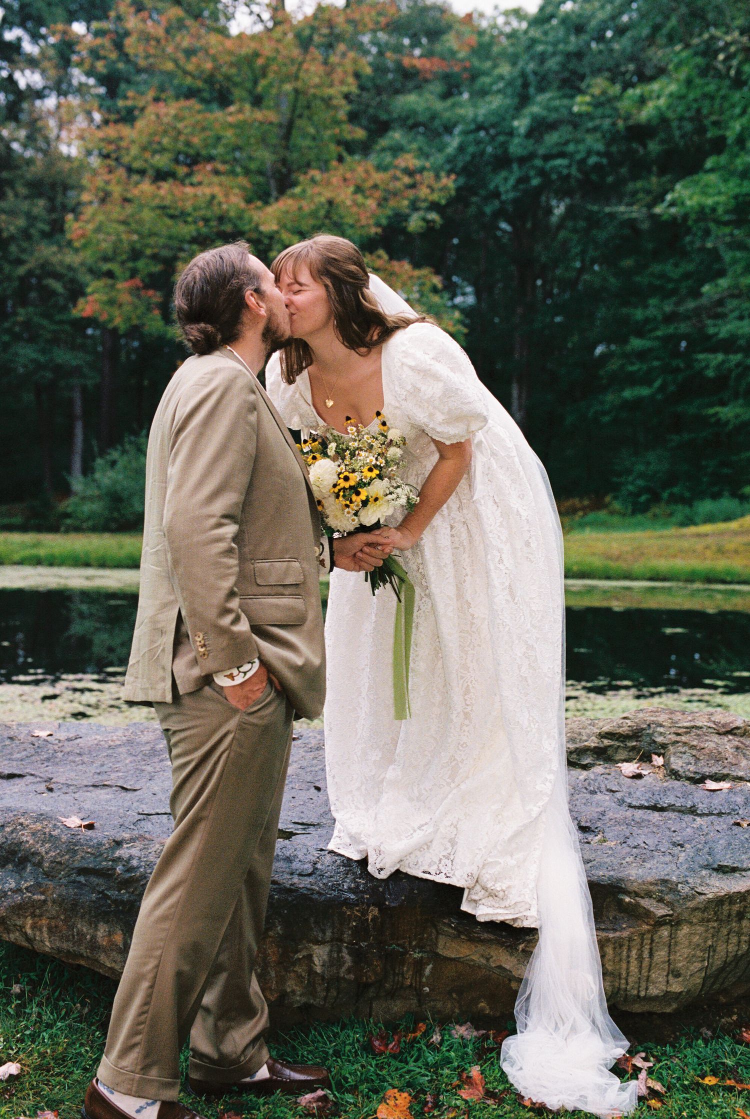 A couple shares their first kiss as newlyweds by a pond in an autumn garden setting with trees in the background.