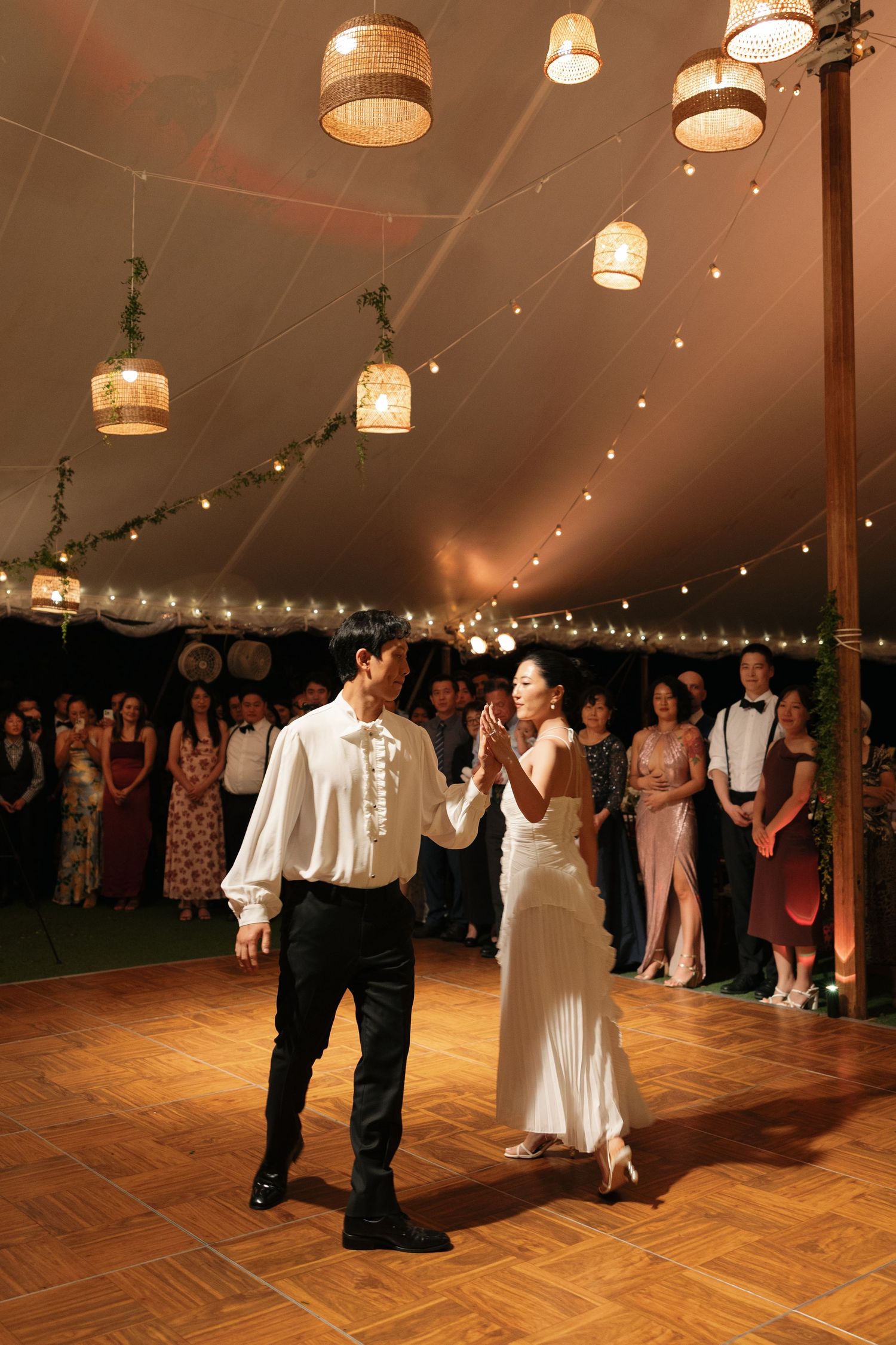 Couple dancing on wooden floor under warm lighting in wedding reception tent with guests watching.