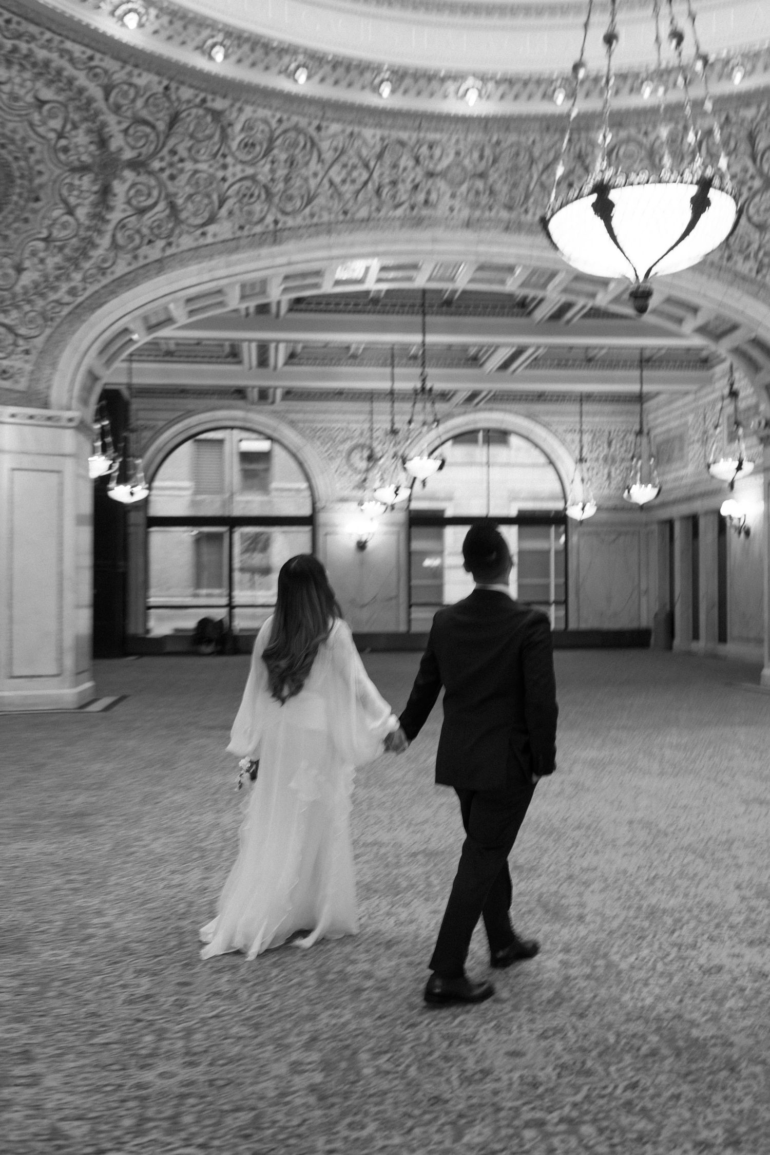 A couple walks hand in hand through an elegant ballroom with ornate ceiling details and vintage hanging light fixtures.