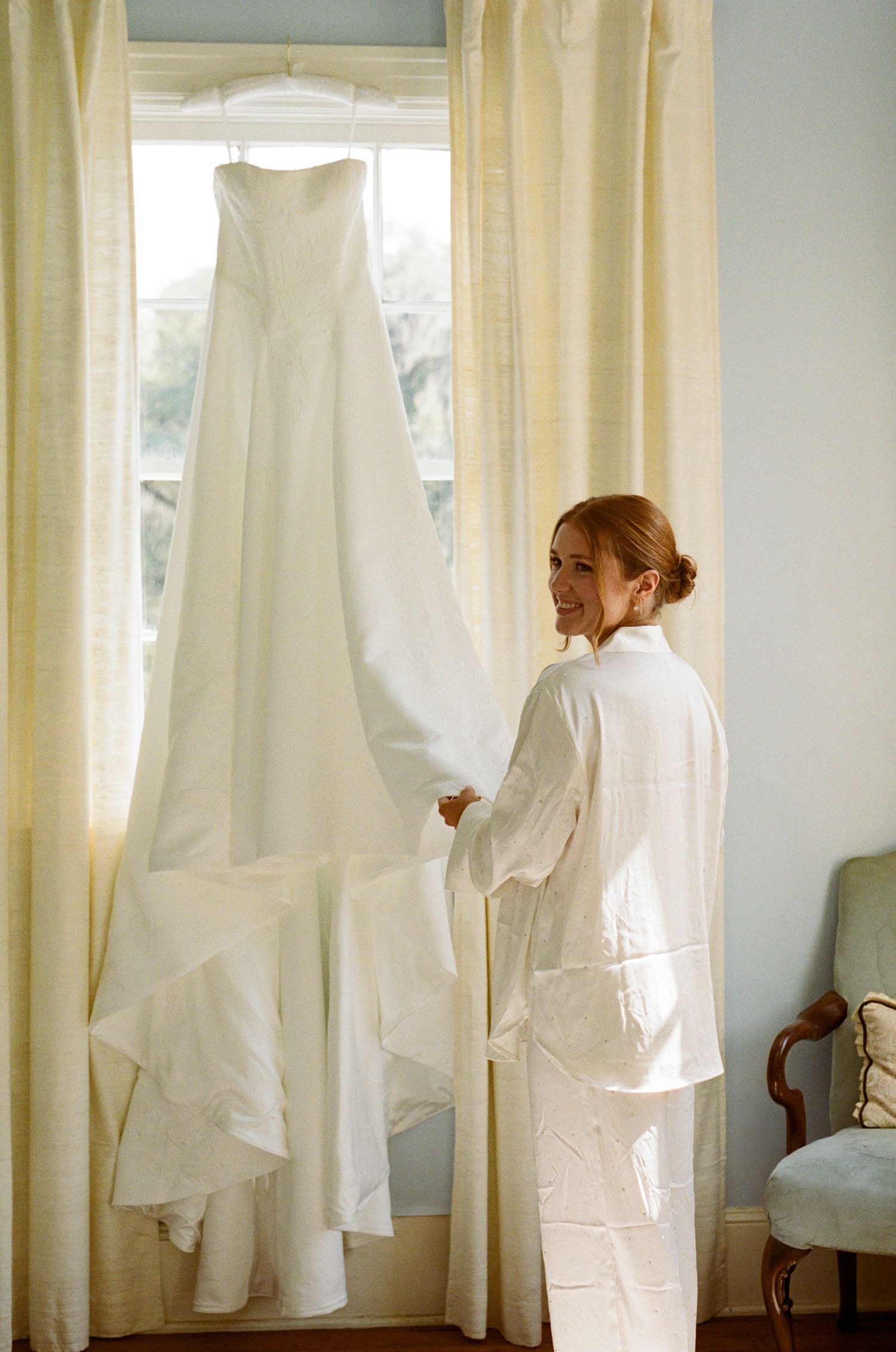 Bride in white silk robe smiling over shoulder at elegant strapless wedding gown hanging by sunlit window.
