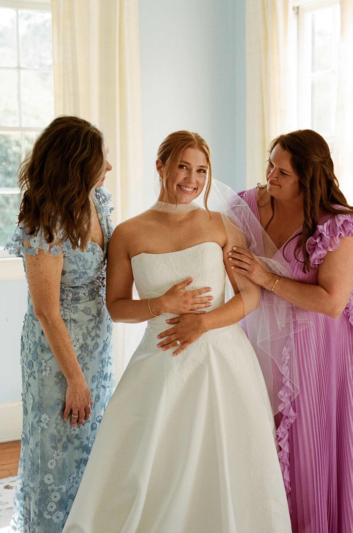 Bride in white strapless gown smiles as two women in blue and lavender dresses help her prepare for her wedding day.