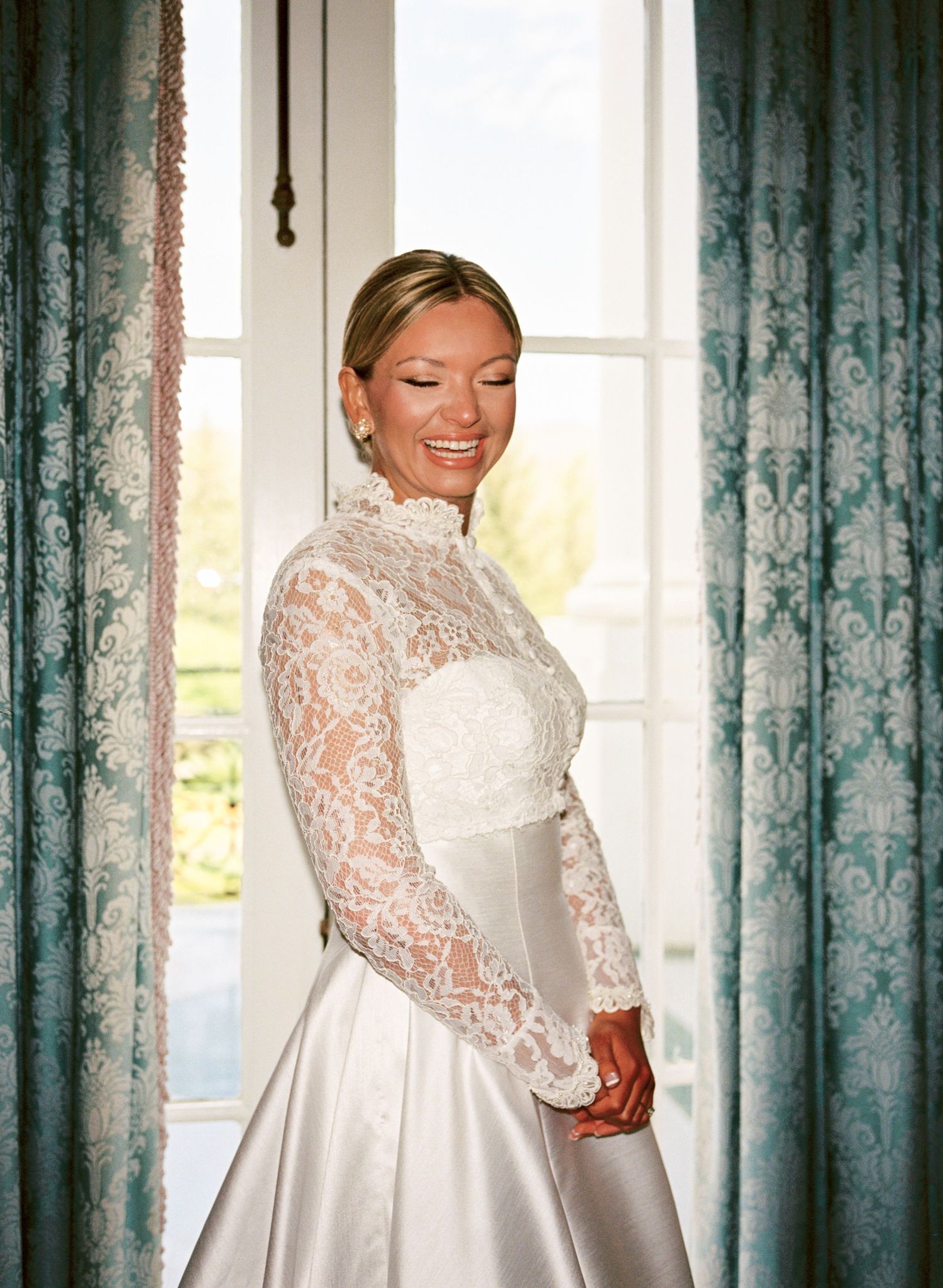 Bride in long-sleeve lace wedding gown smiling by elegant window with teal damask curtains.