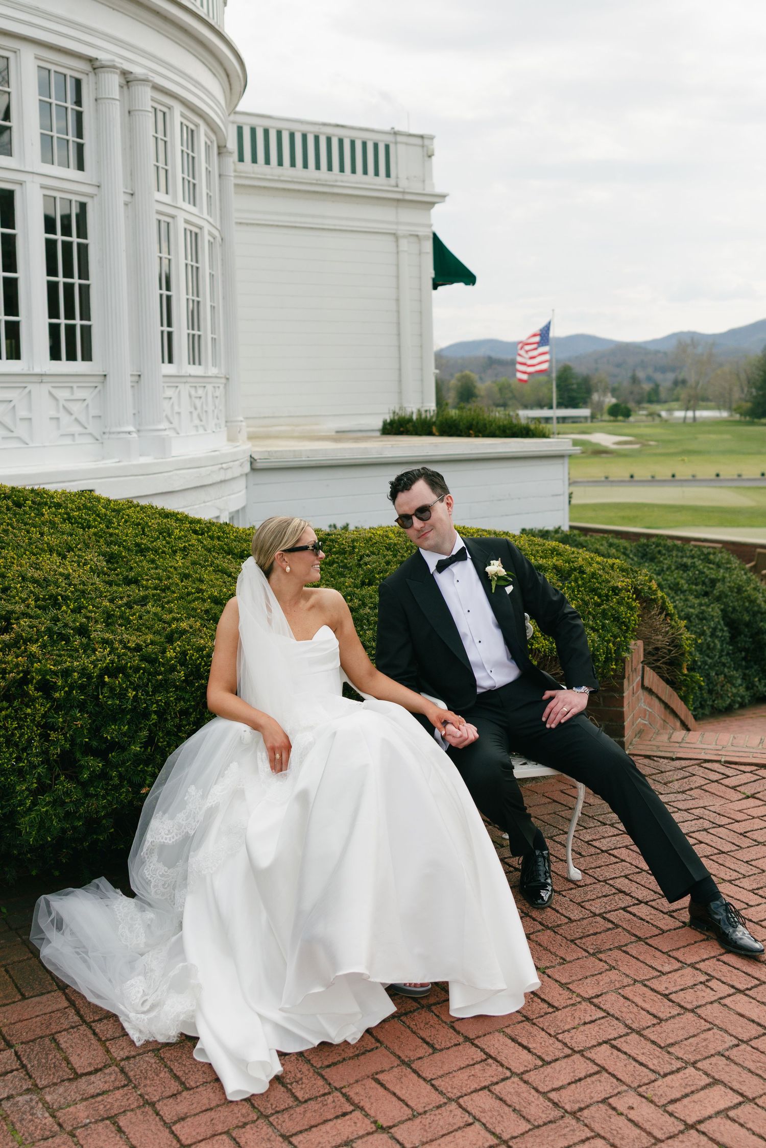 Bride in white gown and groom in black tuxedo sitting together outside a white colonial building with an American flag.