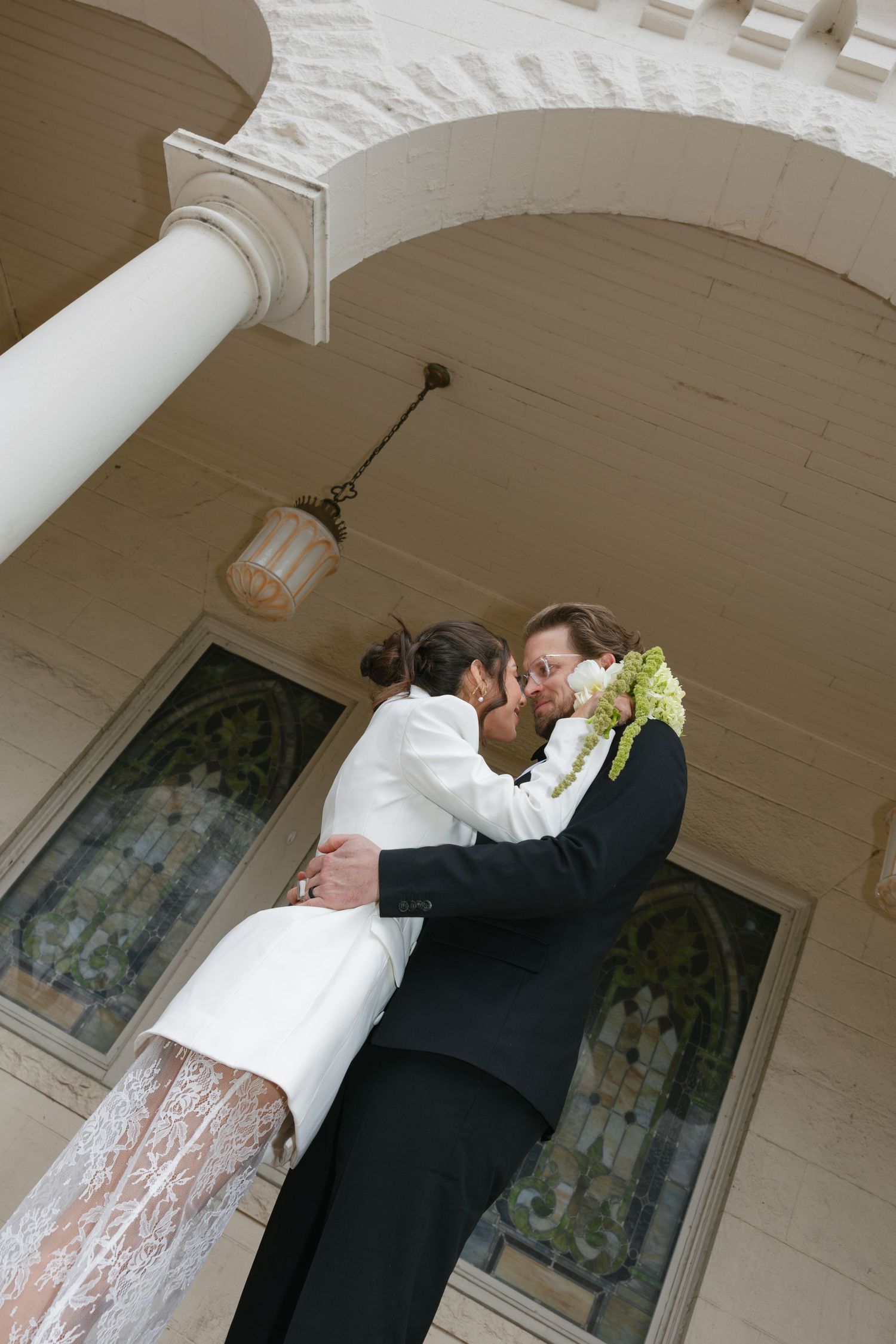 Bride and groom share a romantic kiss under an arched portico with stained glass windows at their wedding venue.