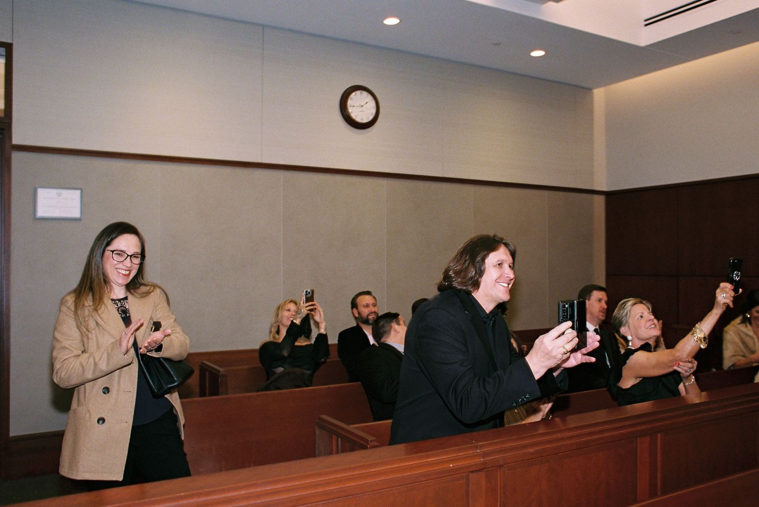 People applauding in a courtroom setting, with wooden benches and a clock on the wall.