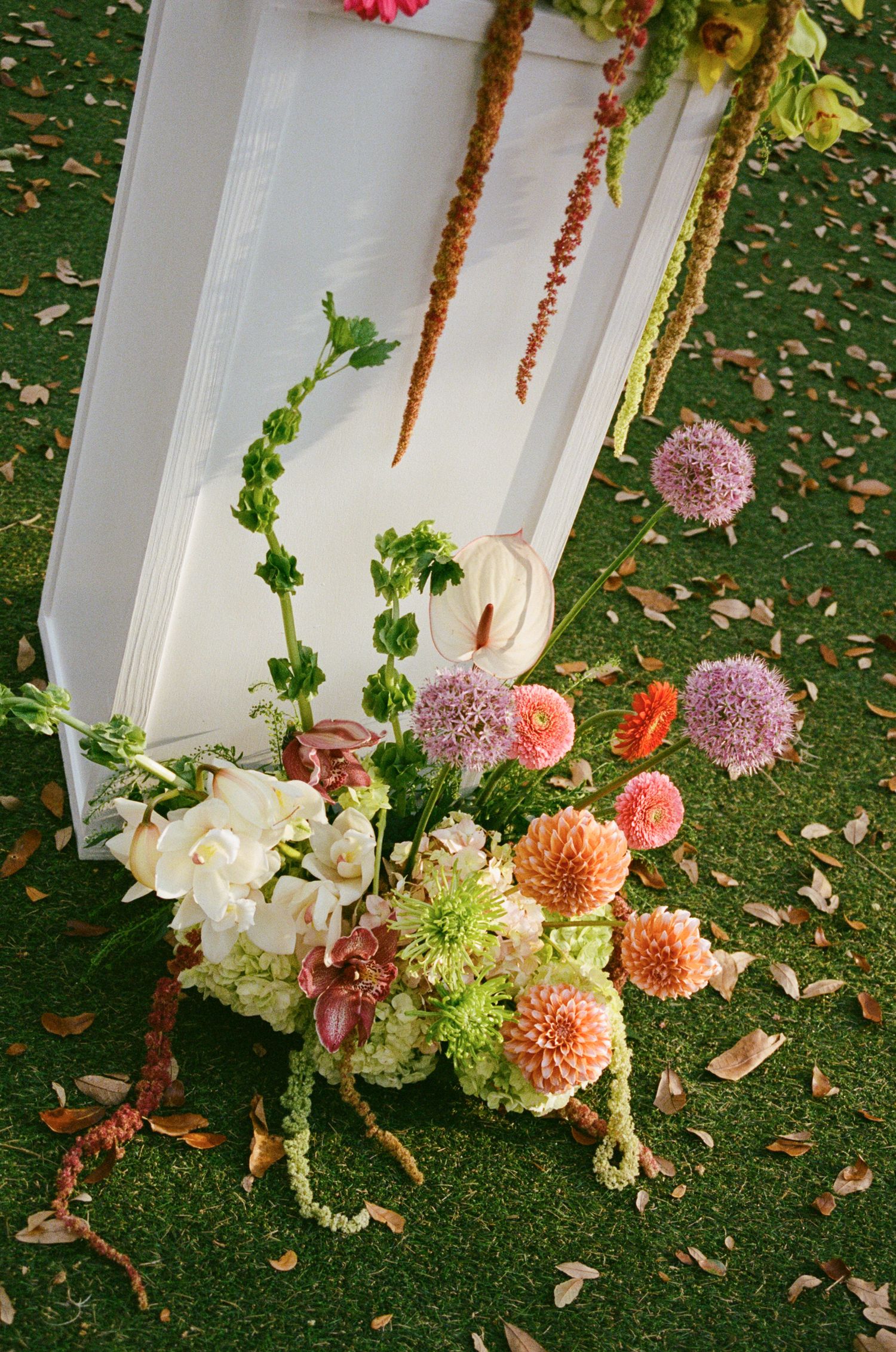 Colorful floral arrangement with dahlias, alliums, and greenery beside a white door frame on grass.