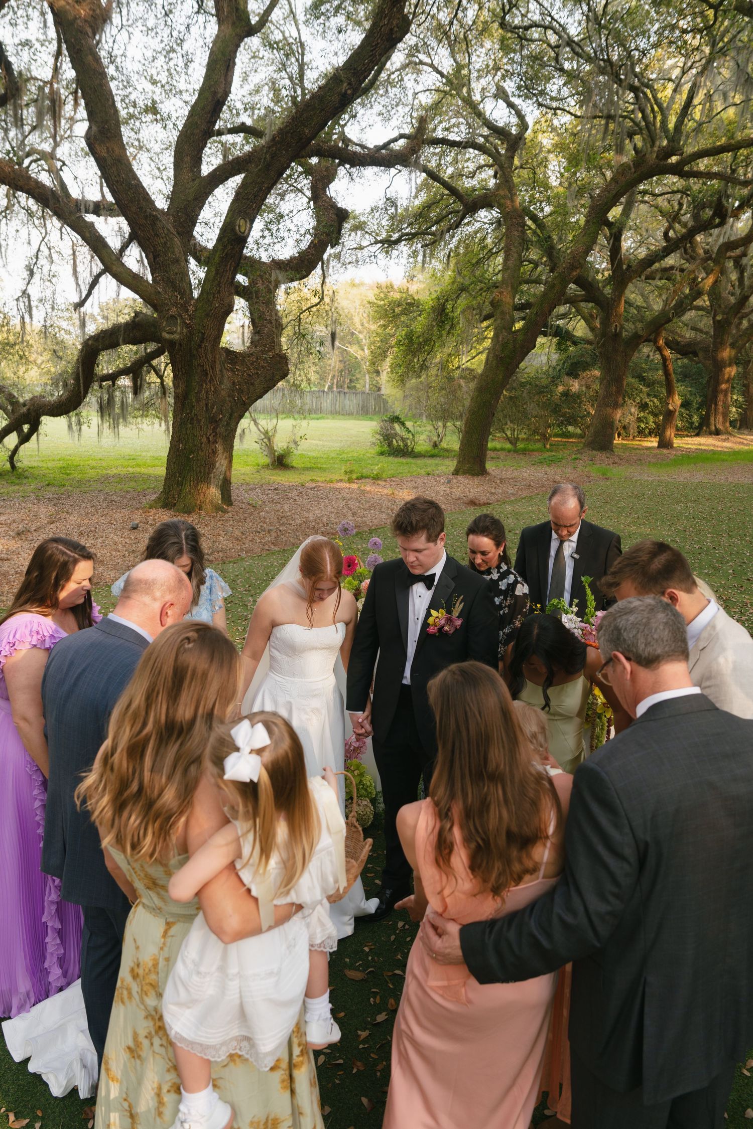 Wedding party holds hands in a circle for an outdoor prayer ceremony under sprawling oak trees.