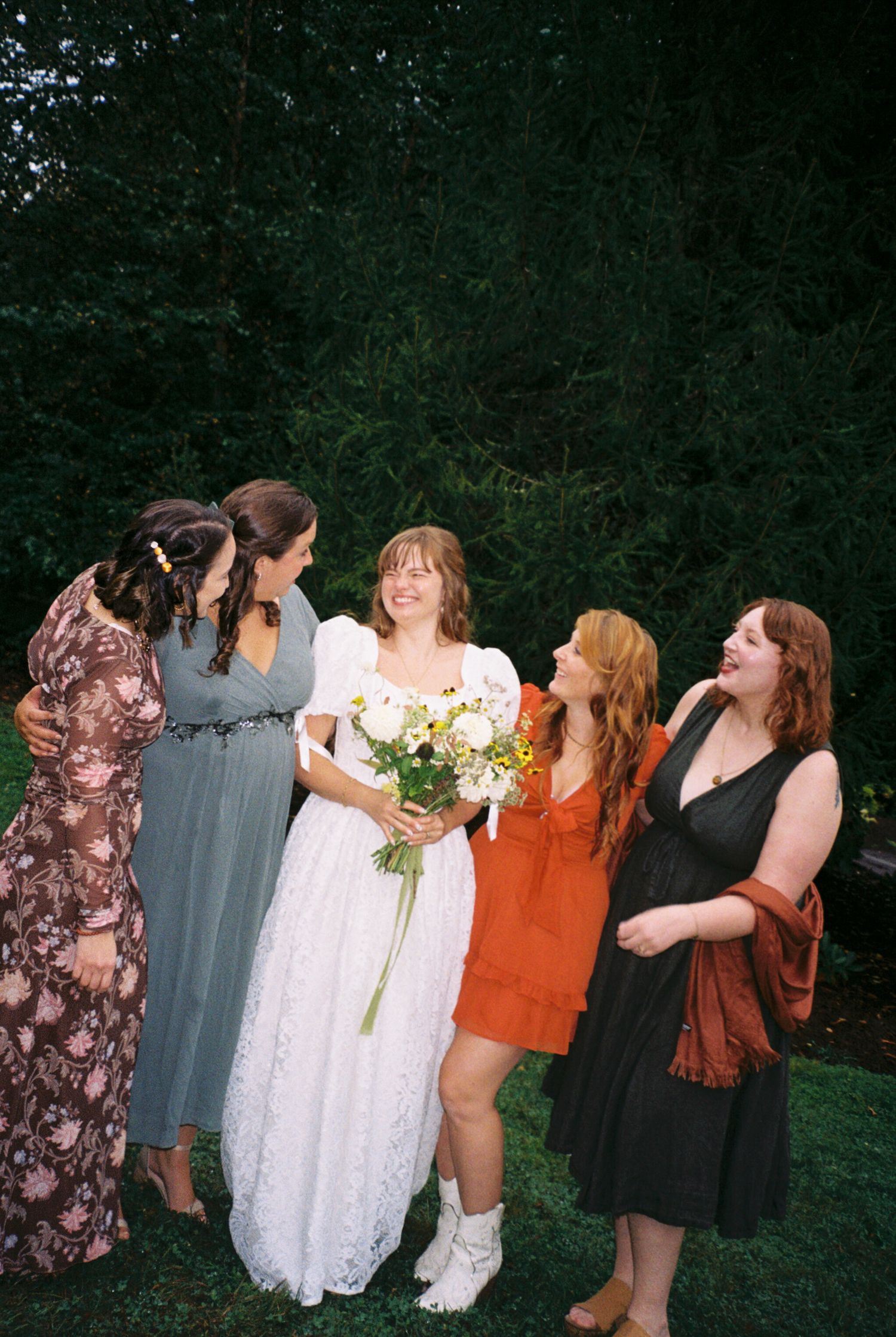 A group of wedding attendees in colorful dresses share laughter and joy while posing together outdoors.