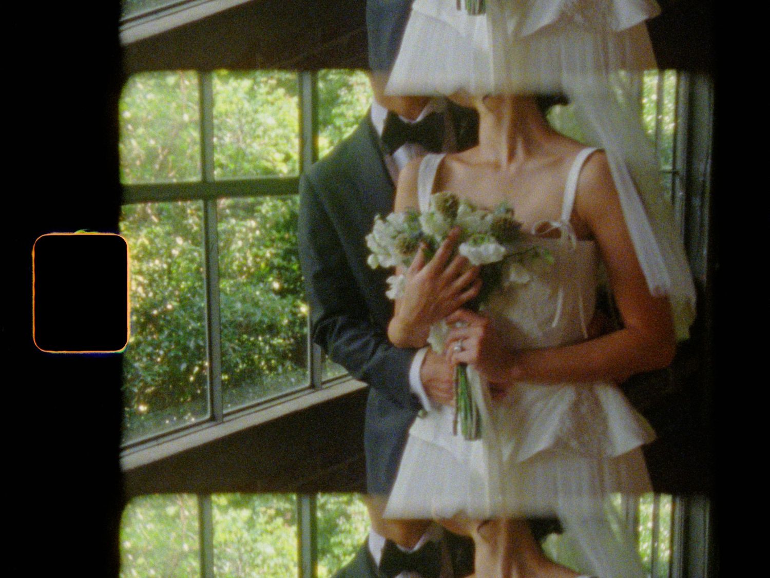 A bride in a white dress and veil holds a bouquet while standing with a groom near a window with green trees outside.