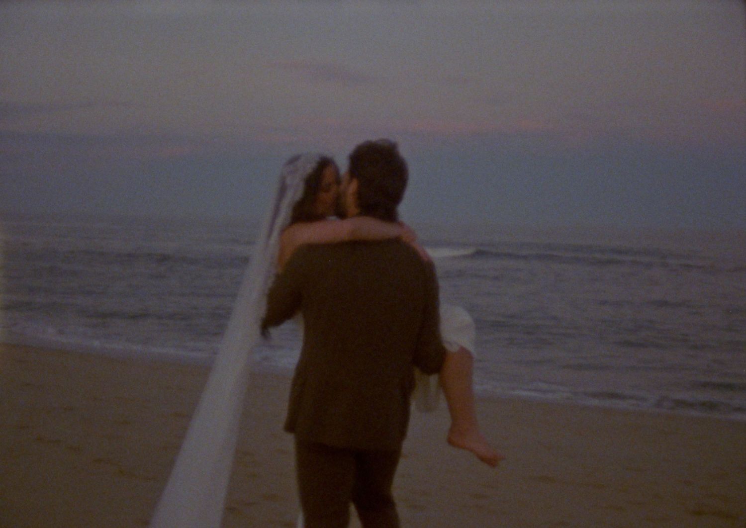 Newlywed couple sharing a romantic kiss on the beach at sunset, bride in white veil and groom in brown suit.