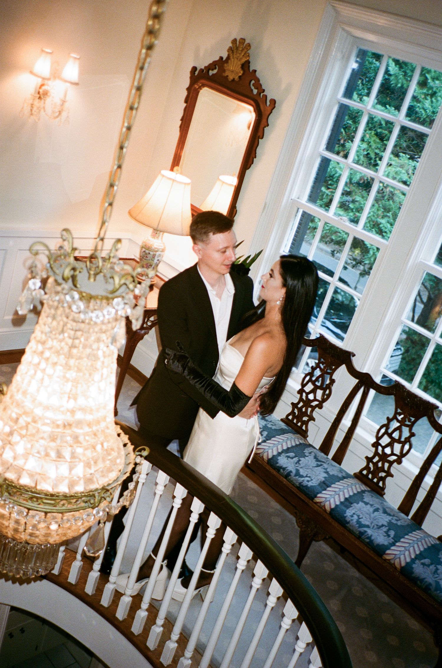 A couple embraces on a curved staircase beside a crystal chandelier in an elegant historic home with tall windows and a carved wooden bench.