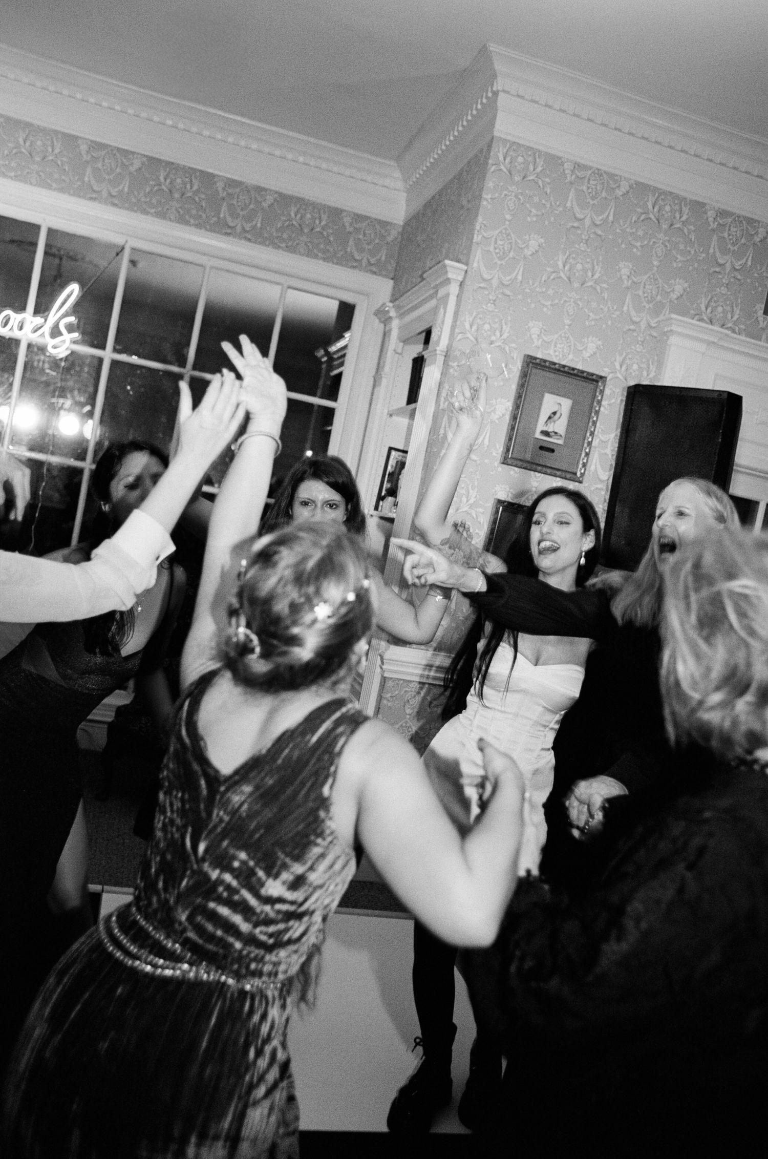 Black and white photo of guests dancing with raised hands and laughing at a lively wedding reception in an elegant room.