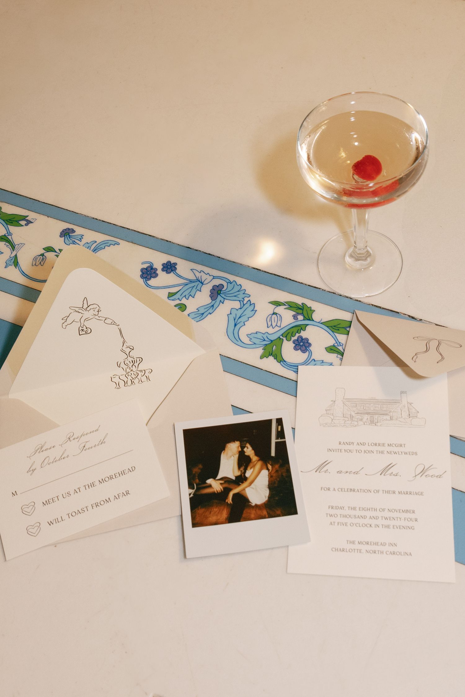 Wedding invitation suite with sketched envelope, Polaroid photo, and blue floral ribbon beside a cocktail topped with a cherry.