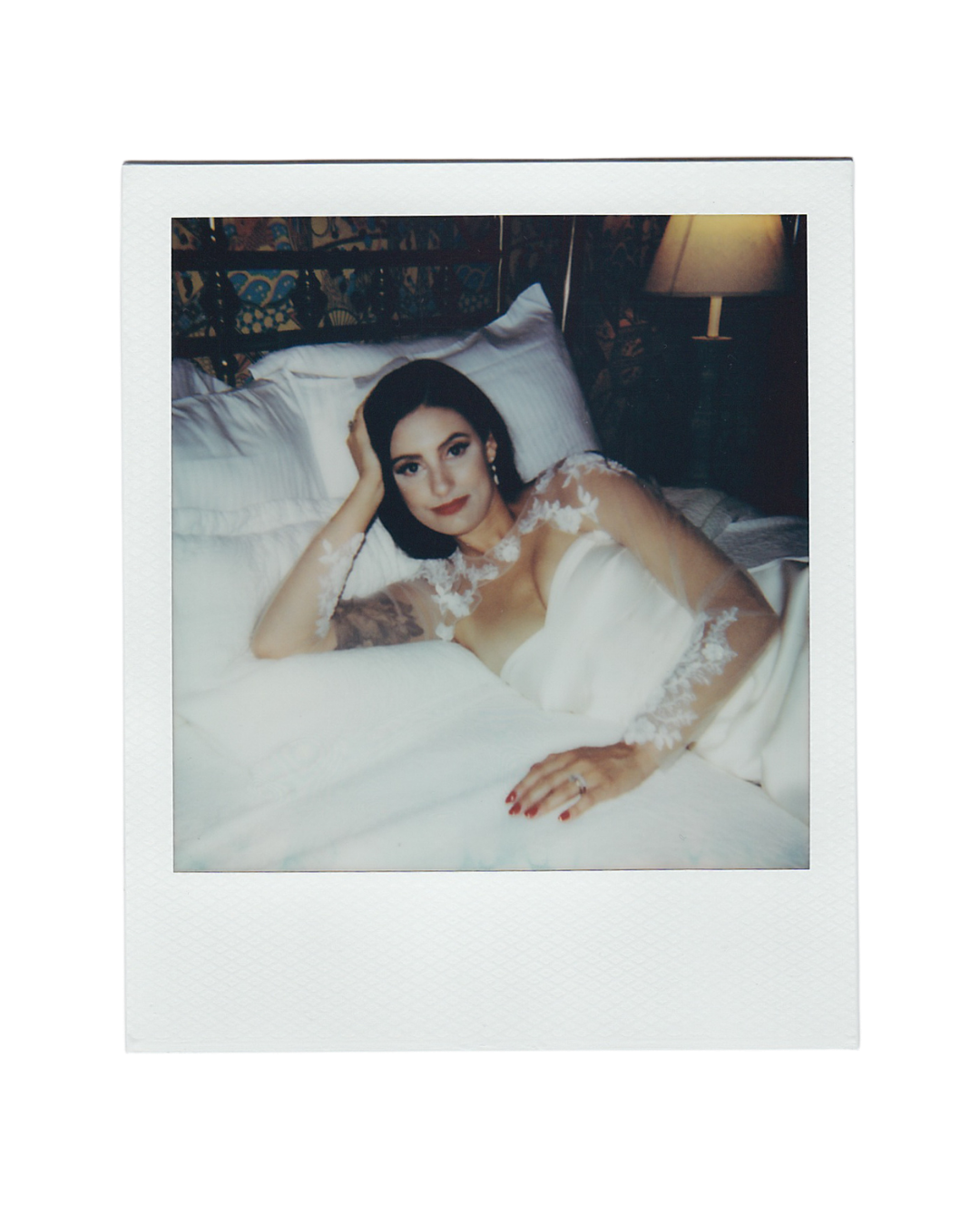 Polaroid-style photo of a woman in a white lace-sleeved bridal gown reclining on white bedding, resting her head on her hand beside a lamp.