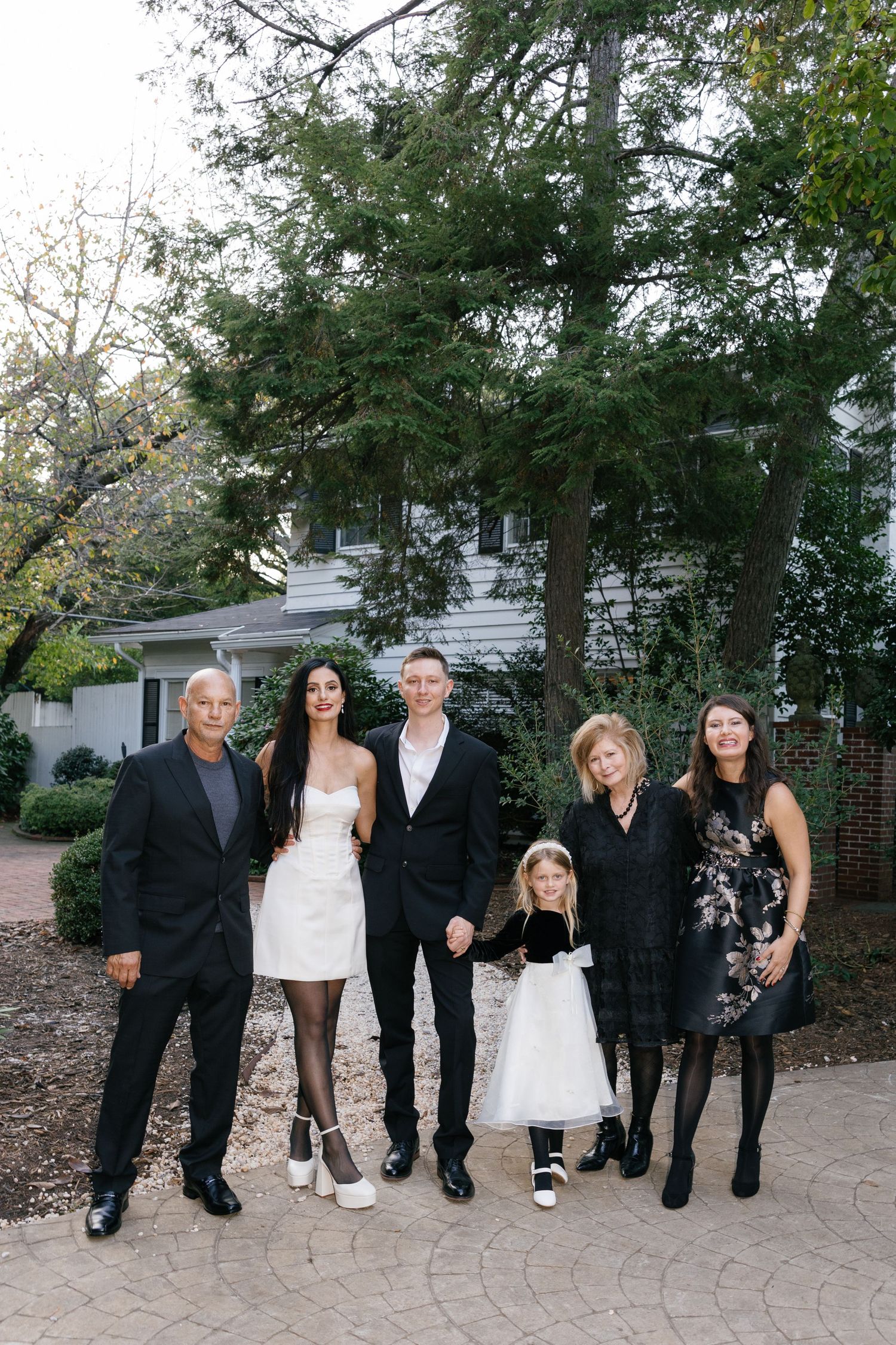 Family of six in black and white formal attire poses on a brick patio in front of a white house and tall evergreen trees.