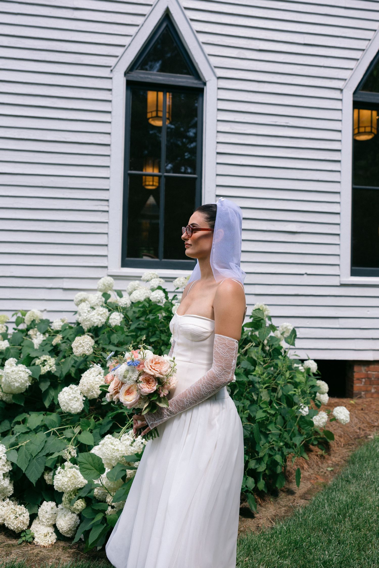 A bride in a white strapless gown, sheer gloves and short veil holds a peach bouquet beside white hydrangeas and a white clapboard building.