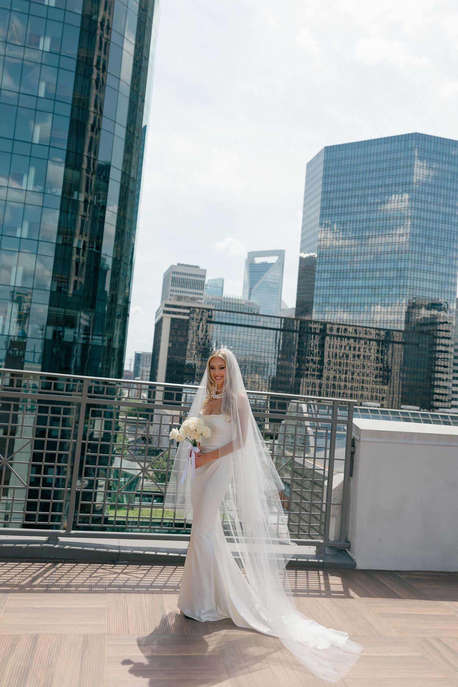 Elegant bride with cathedral veil stands against modern city architecture.