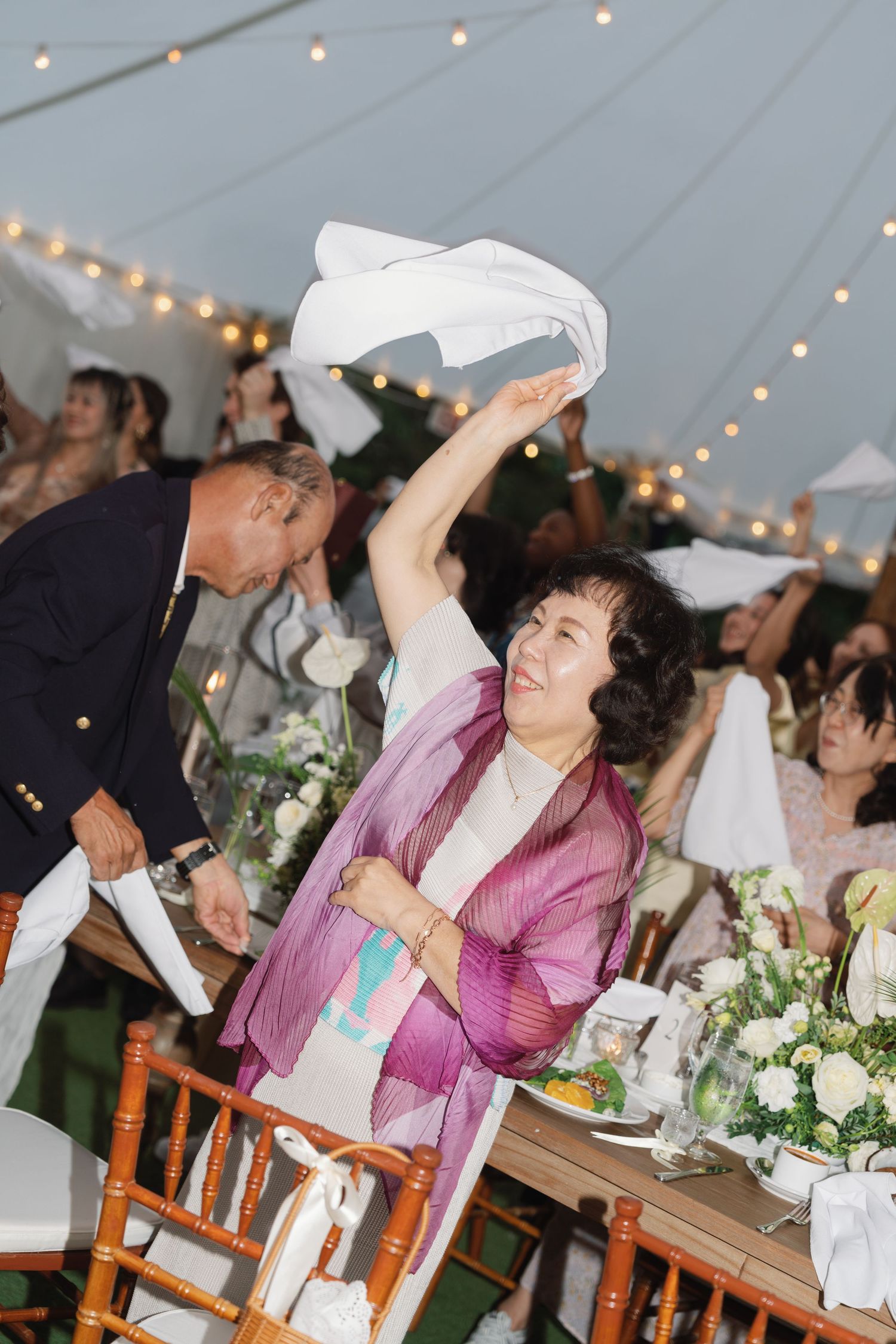 Wedding guests share a playful moment at a reception under a tent decorated with string lights and floral centerpieces.