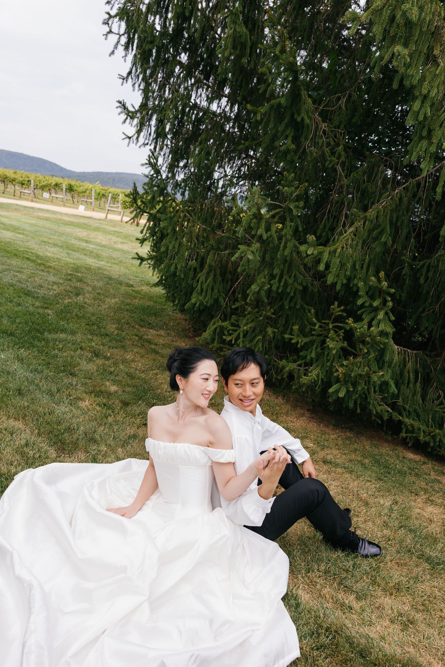 A couple in wedding attire rests together on grassy hillside beneath evergreen trees.