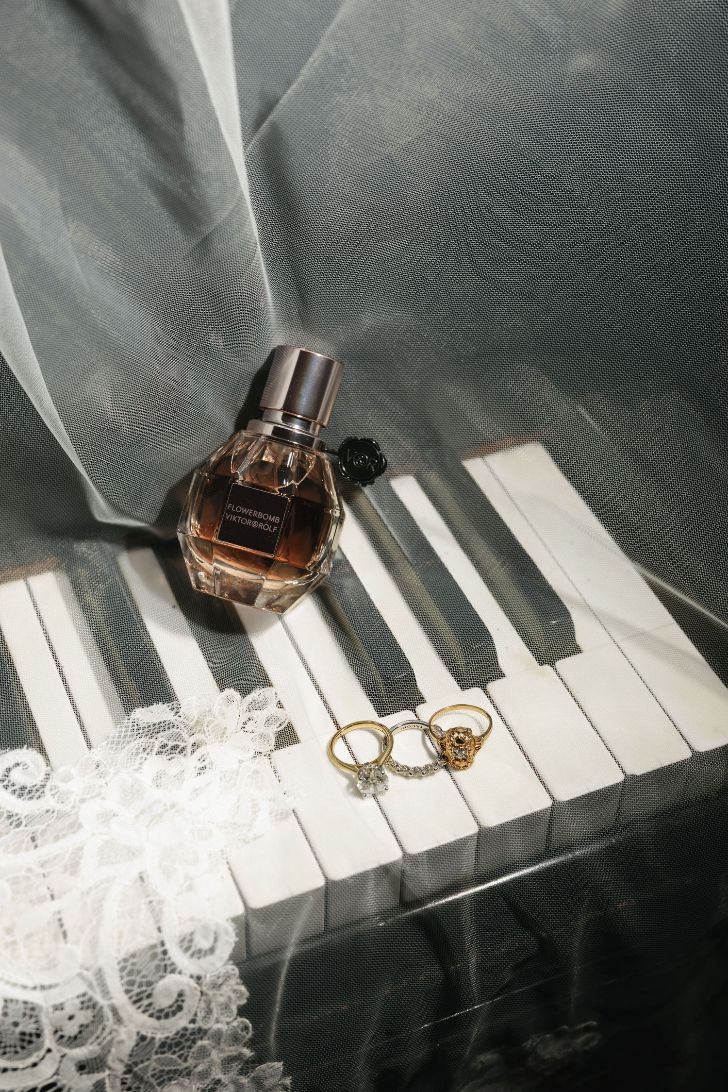 Perfume bottle and gold rings on piano keys with white lace and sheer fabric, elegant bridal flat lay.