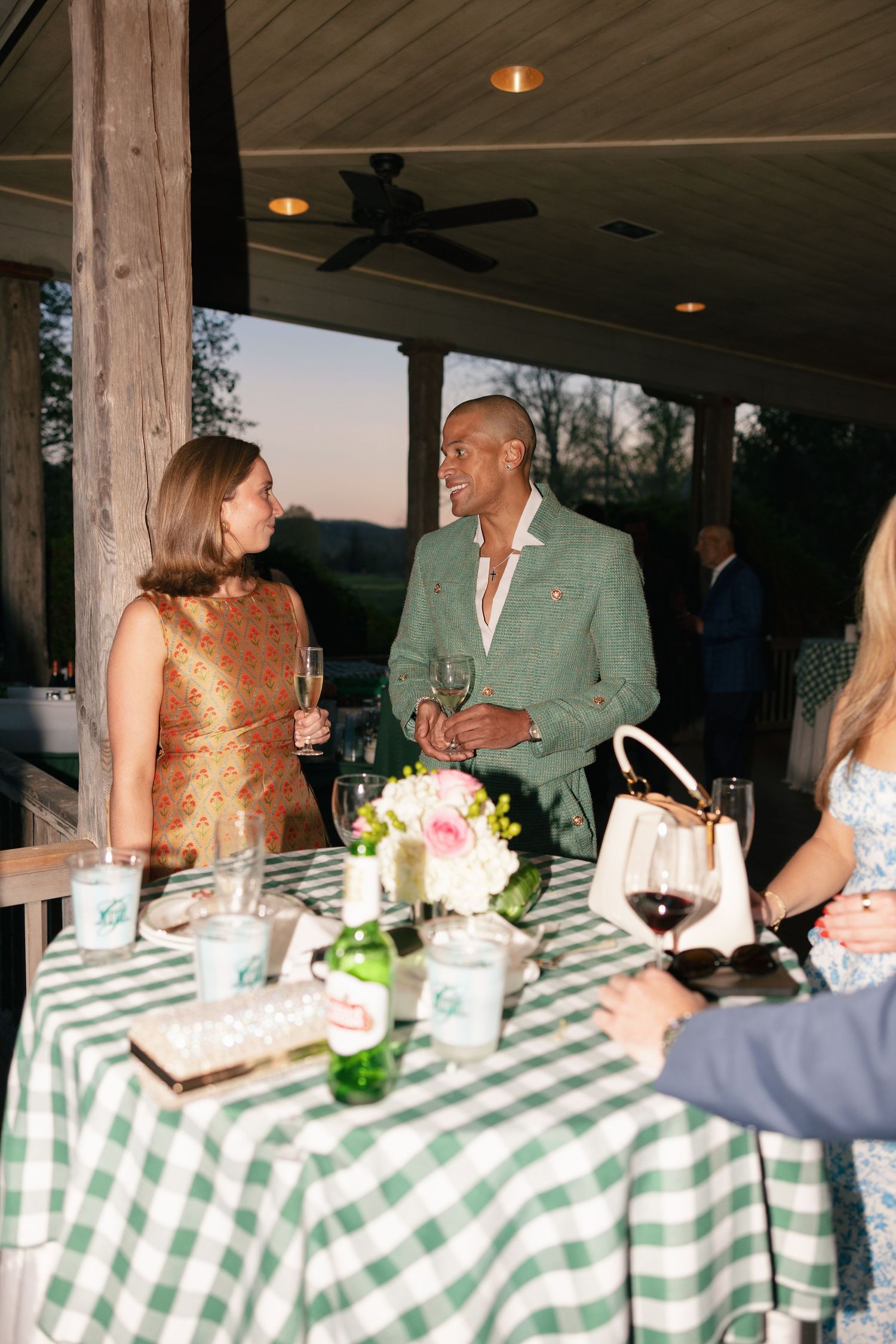 Two guests in elegant attire chat over wine at a green checkered table during an outdoor evening event.