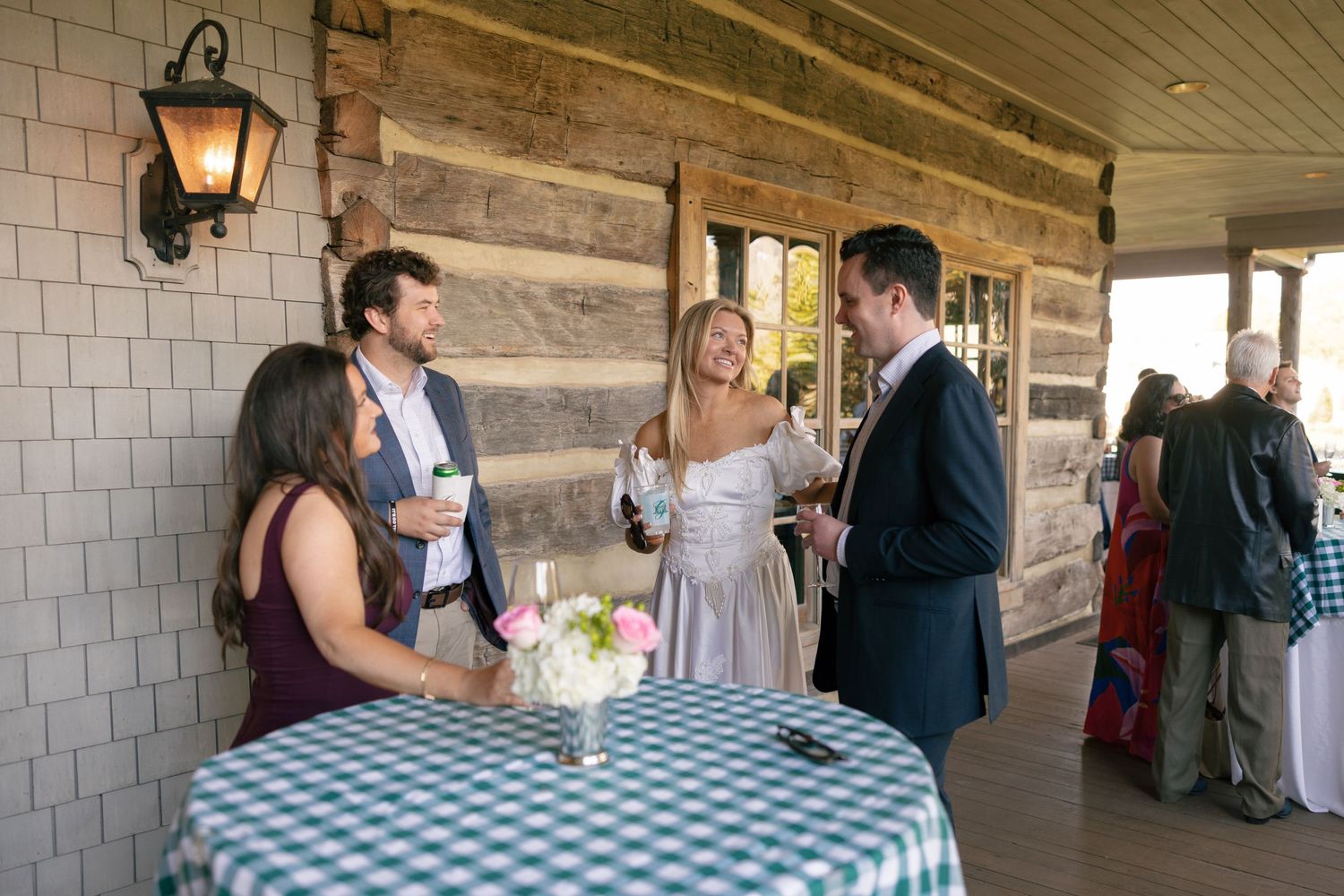 Four guests socializing at a rustic outdoor patio reception, holding drinks near a gingham-covered cocktail table.