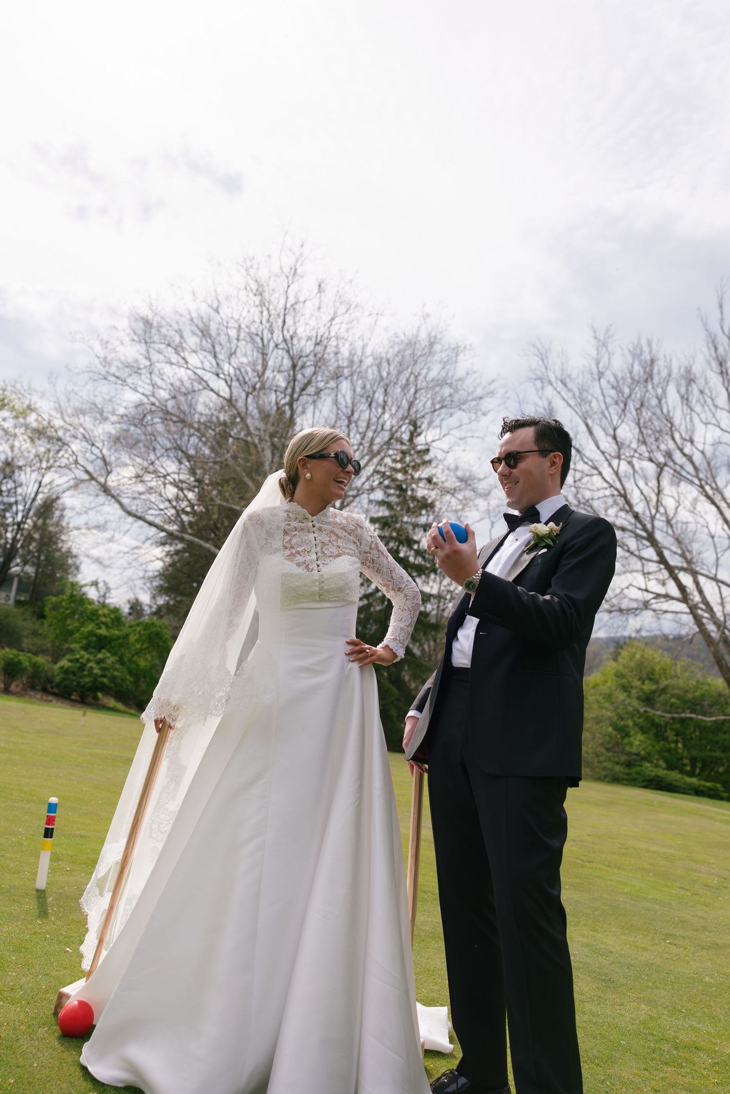 Bride in white lace gown and groom in black tuxedo playing croquet on green lawn at outdoor wedding.