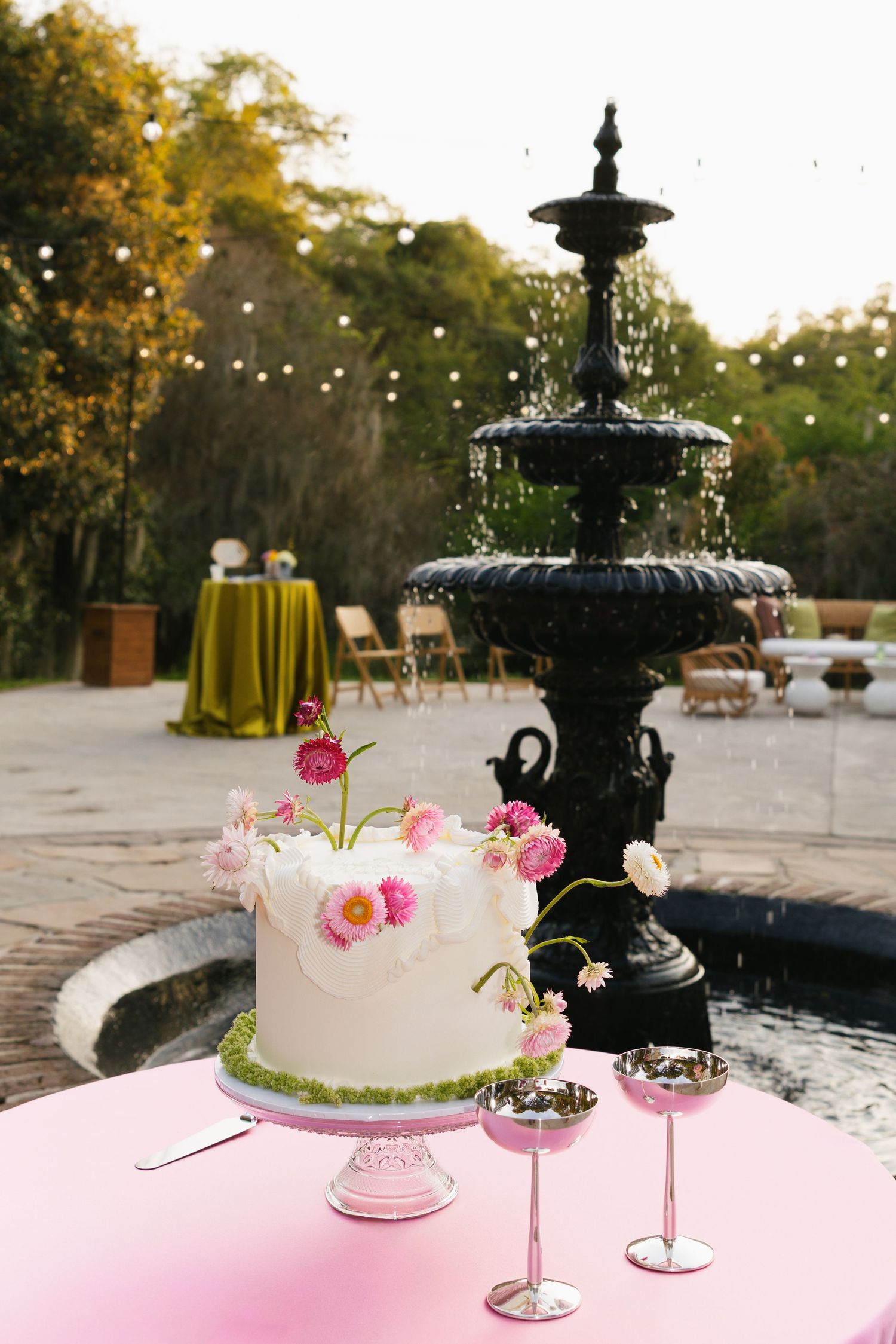 White wedding cake with pink flowers on a pink table beside a black fountain at an outdoor garden reception.