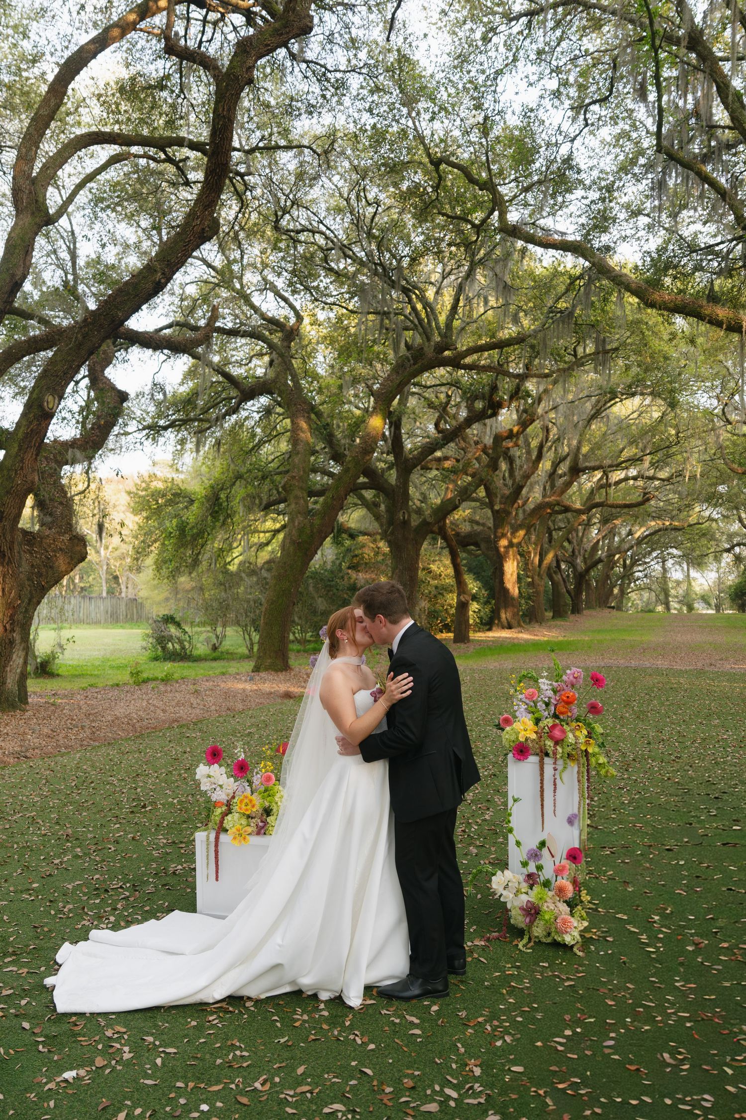 Bride and groom share first kiss outdoors under sprawling oak trees, flanked by colorful floral arrangements on white pedestals.