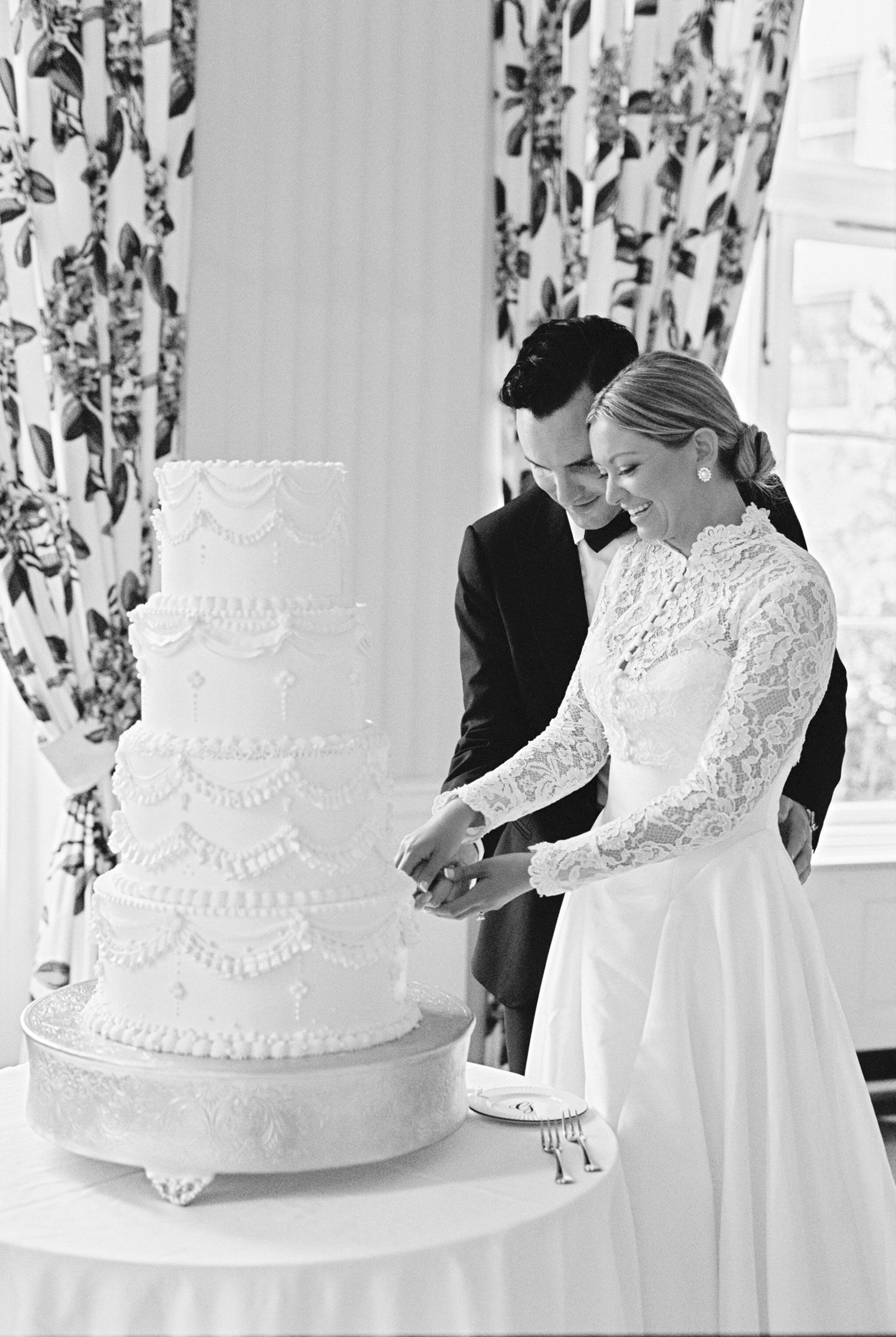 Black and white photo of a bride and groom cutting a tall four-tier wedding cake at their wedding reception.