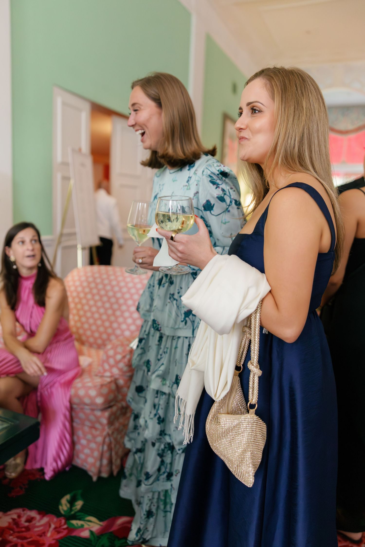 Two elegantly dressed women holding white wine glasses at a formal event in a green-walled venue.
