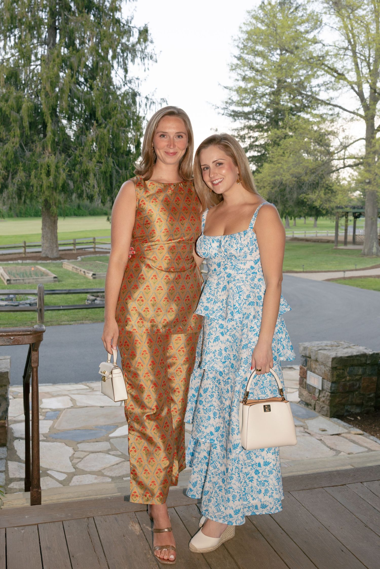 Two women in elegant floral and gold maxi dresses pose outdoors at a garden event holding white handbags.