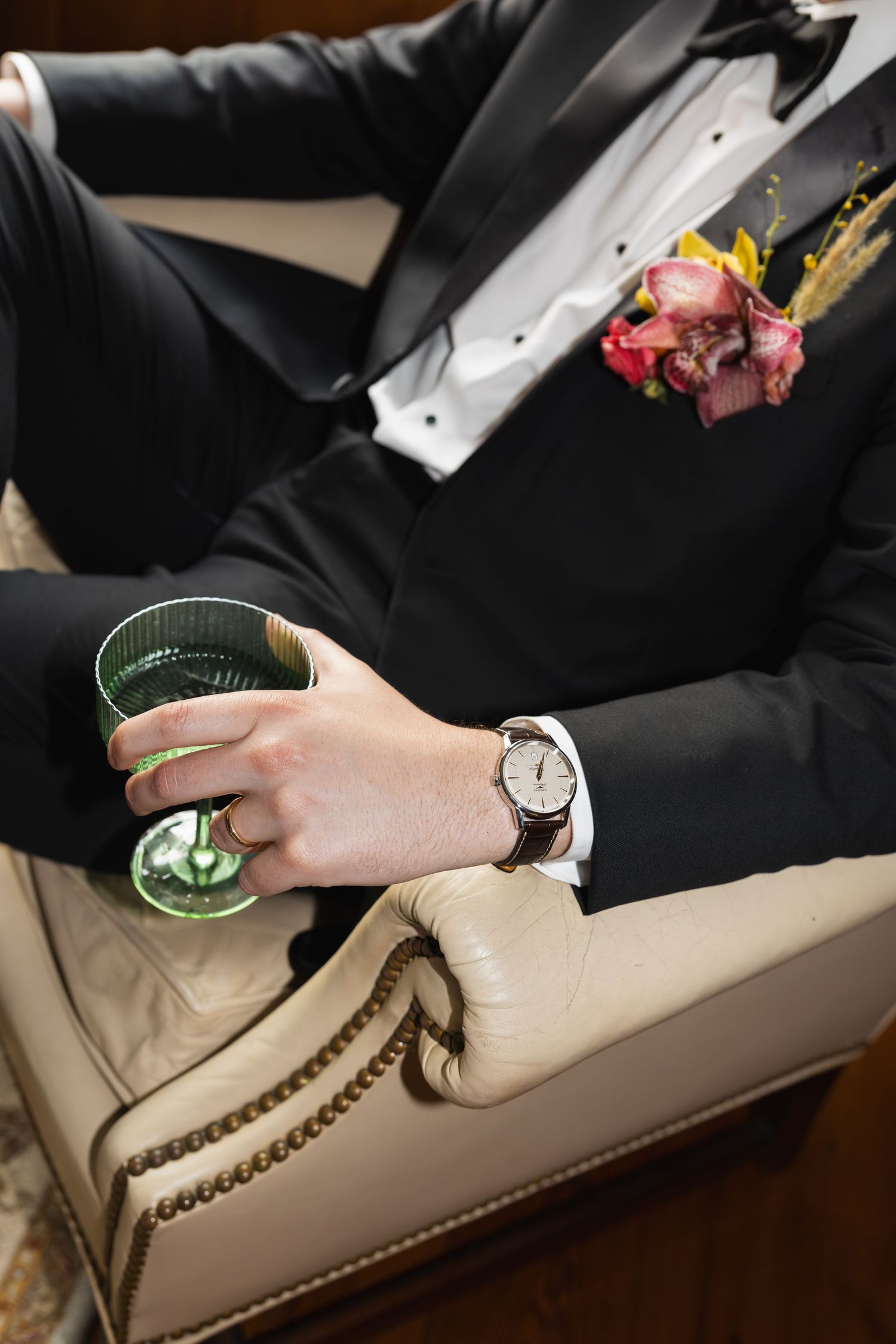 Man in tuxedo with floral boutonniere holding green cocktail glass, wearing brown leather watch, seated on cream chair.