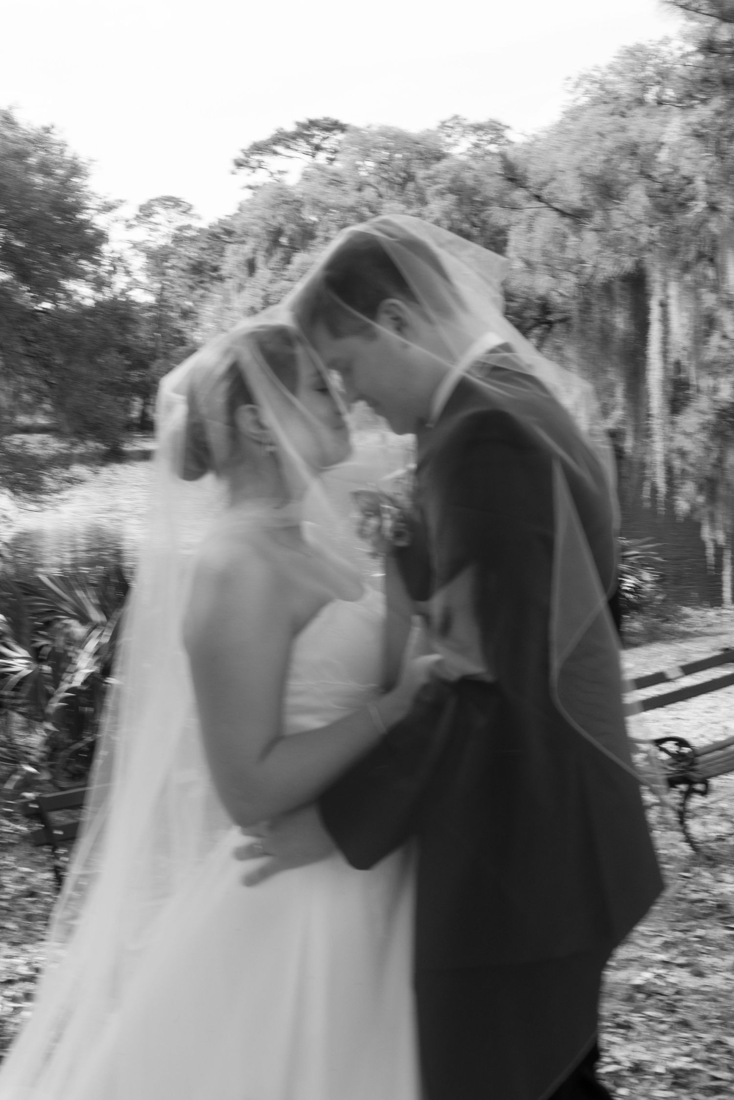 Black and white photo of a bride and groom sharing a romantic moment under a wedding veil outdoors.