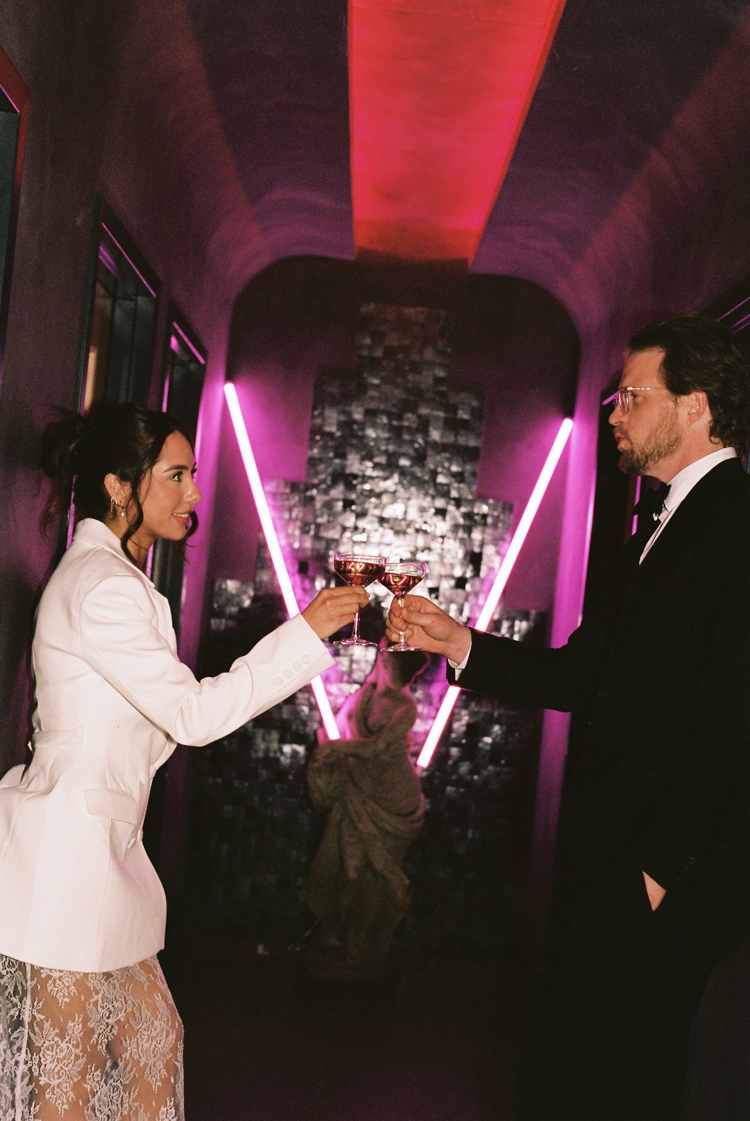 Couple in elegant attire toasting with wine glasses in a moody corridor with pink neon lights and decorative backdrop.