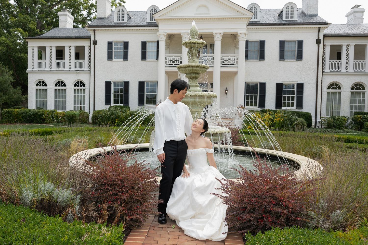 A couple in wedding attire walks together on a winding garden path in front of a grand white colonial mansion.