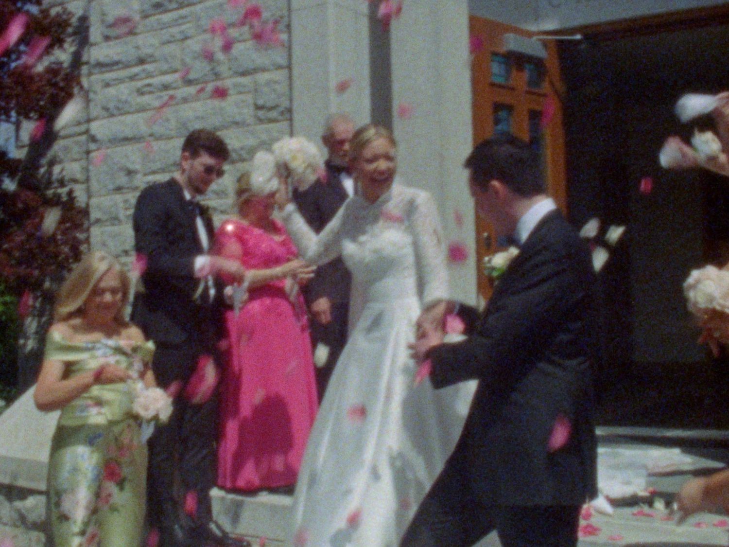 Bride and groom exit church surrounded by wedding guests tossing rose petals in a joyful outdoor ceremony.