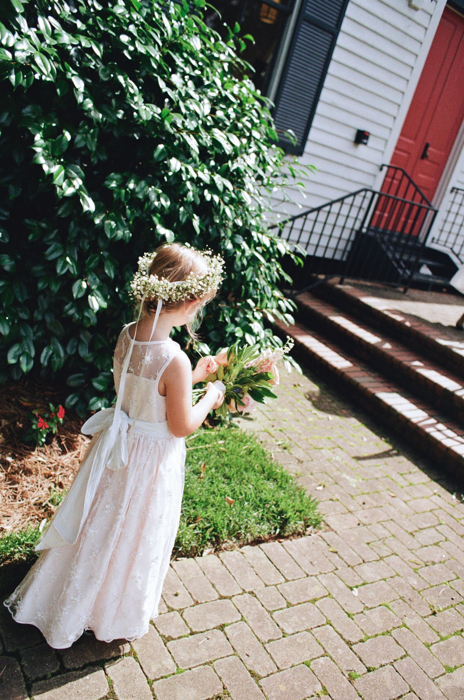 Flower girl in a white dress and floral crown holding a bouquet while walking along a brick path beside green shrubs and a red door.