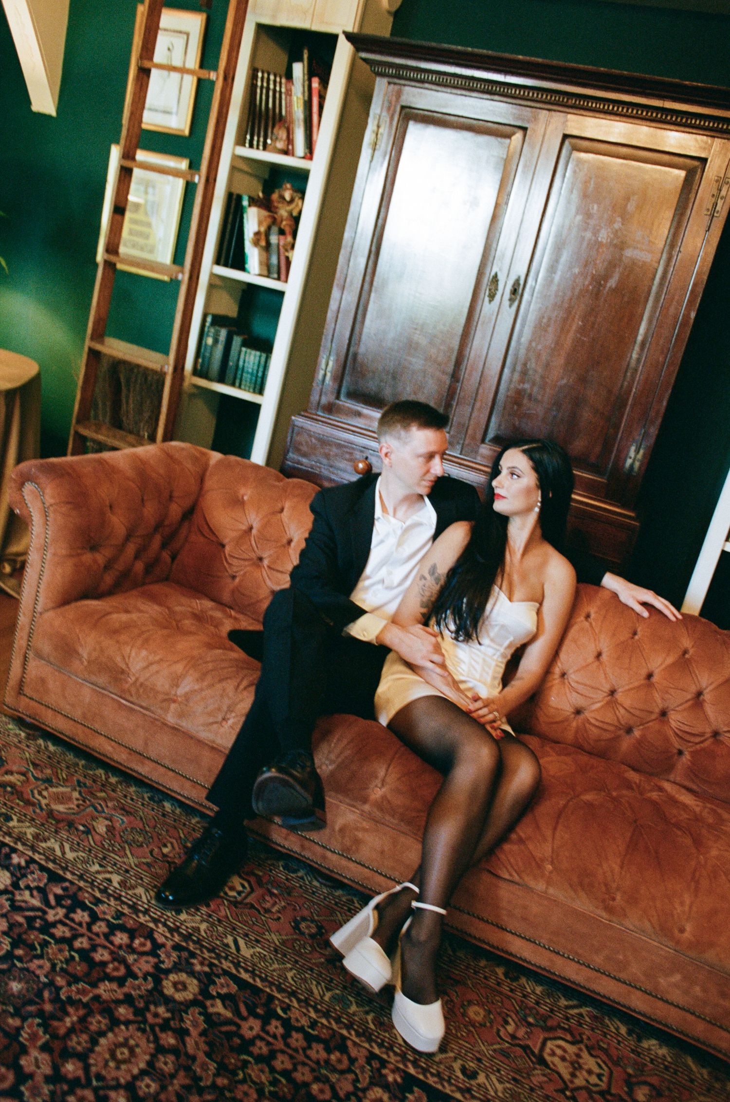 Couple posing together on a tufted brown leather sofa in a cozy room with green walls, an antique wooden armoire, and a patterned rug.