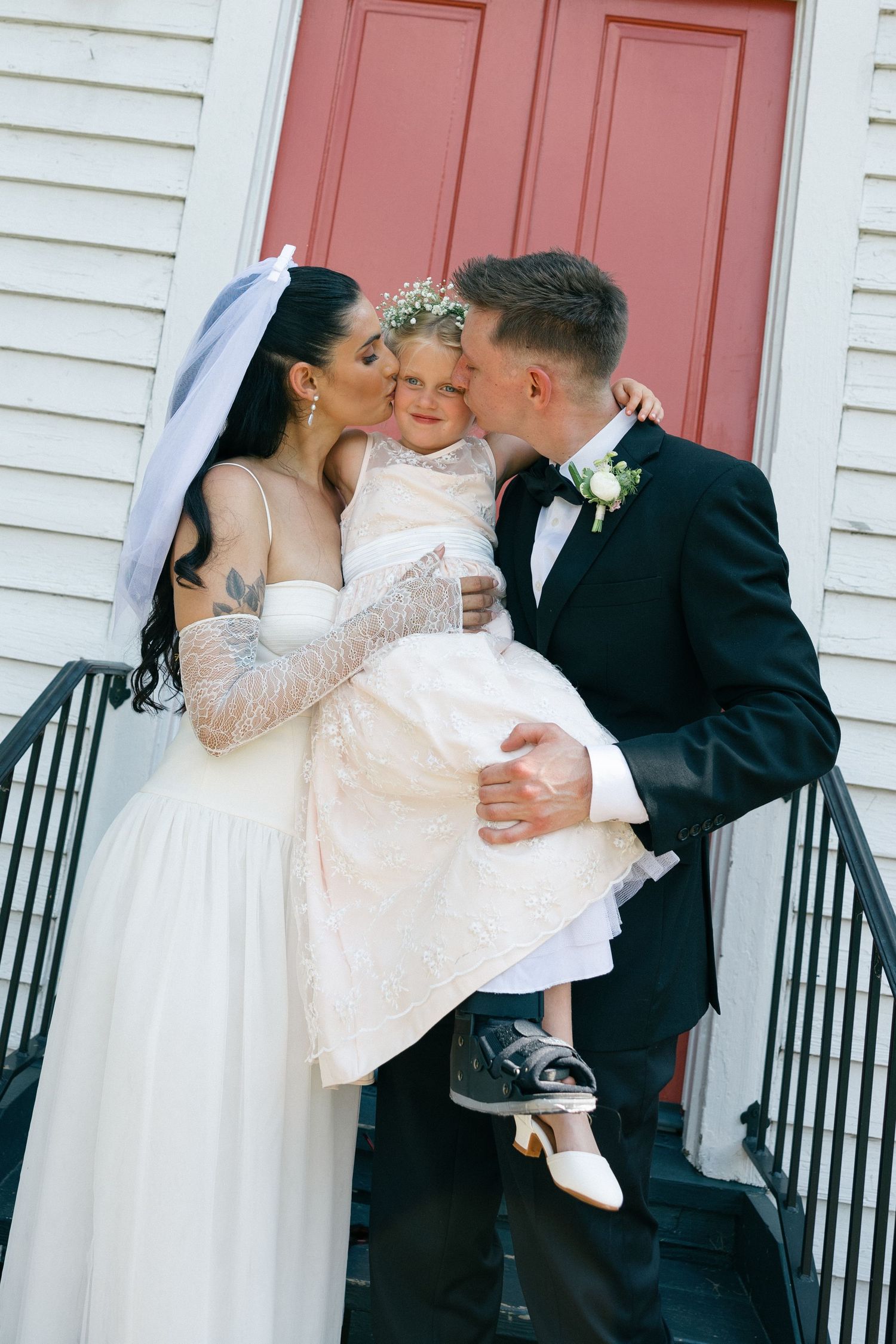 Bride and groom kiss their young flower girl on the cheek while holding her on the steps of a white building with a red door.