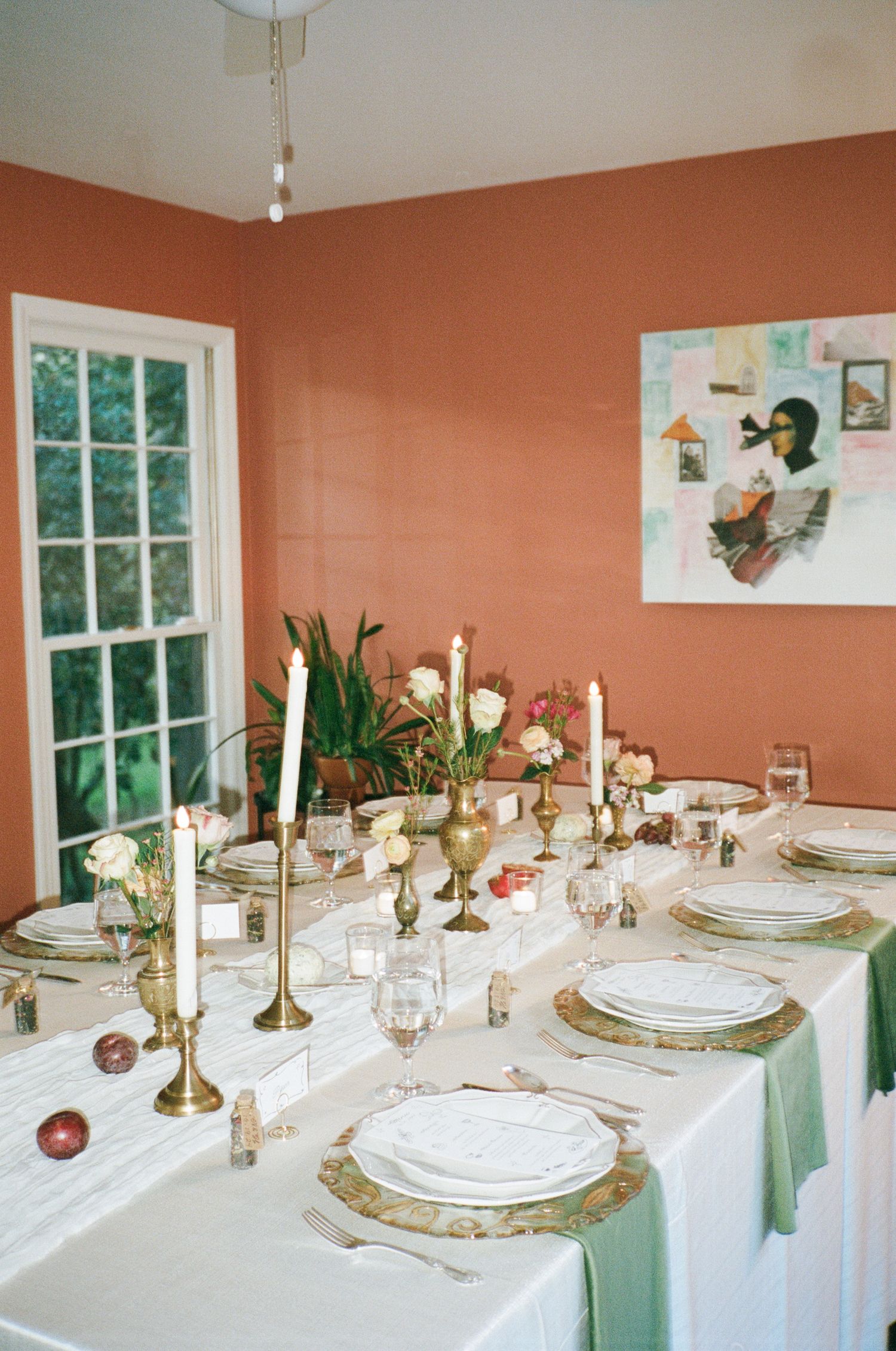 Elegantly set dining table with white linens, green napkins, brass candlesticks, fresh flowers and plates in a coral-walled room.
