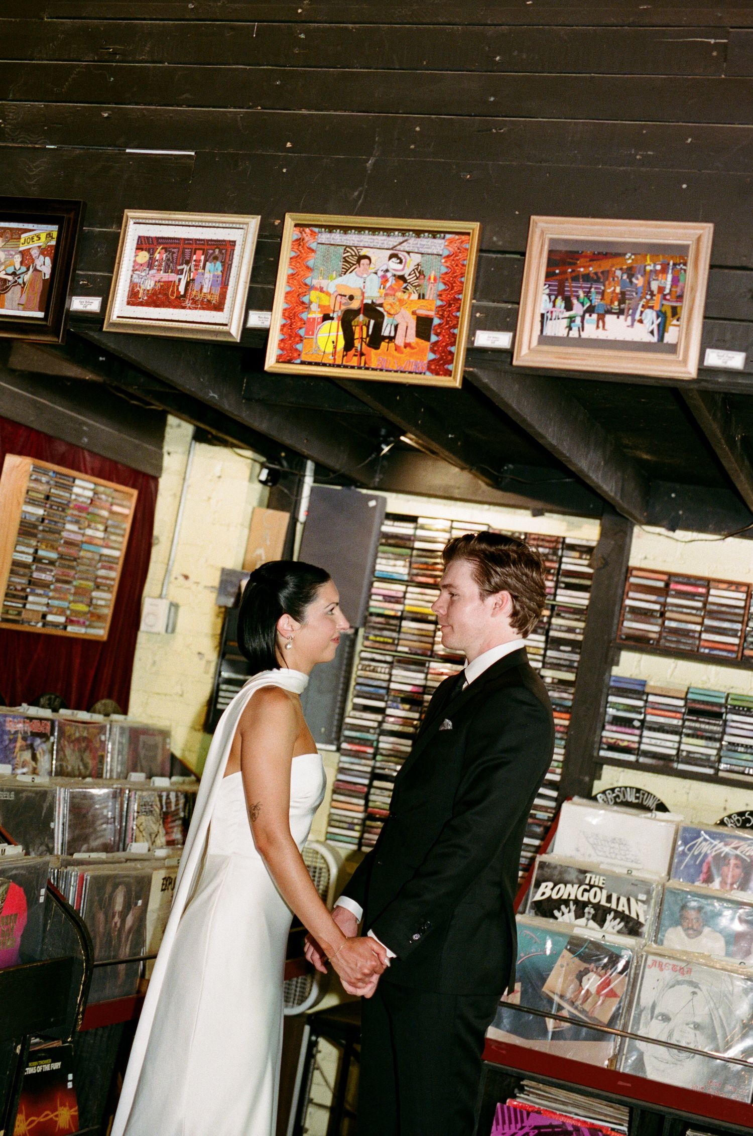 Bride in a white gown and groom in a dark suit hold hands during a wedding ceremony inside a record store with framed art and CD shelves.
