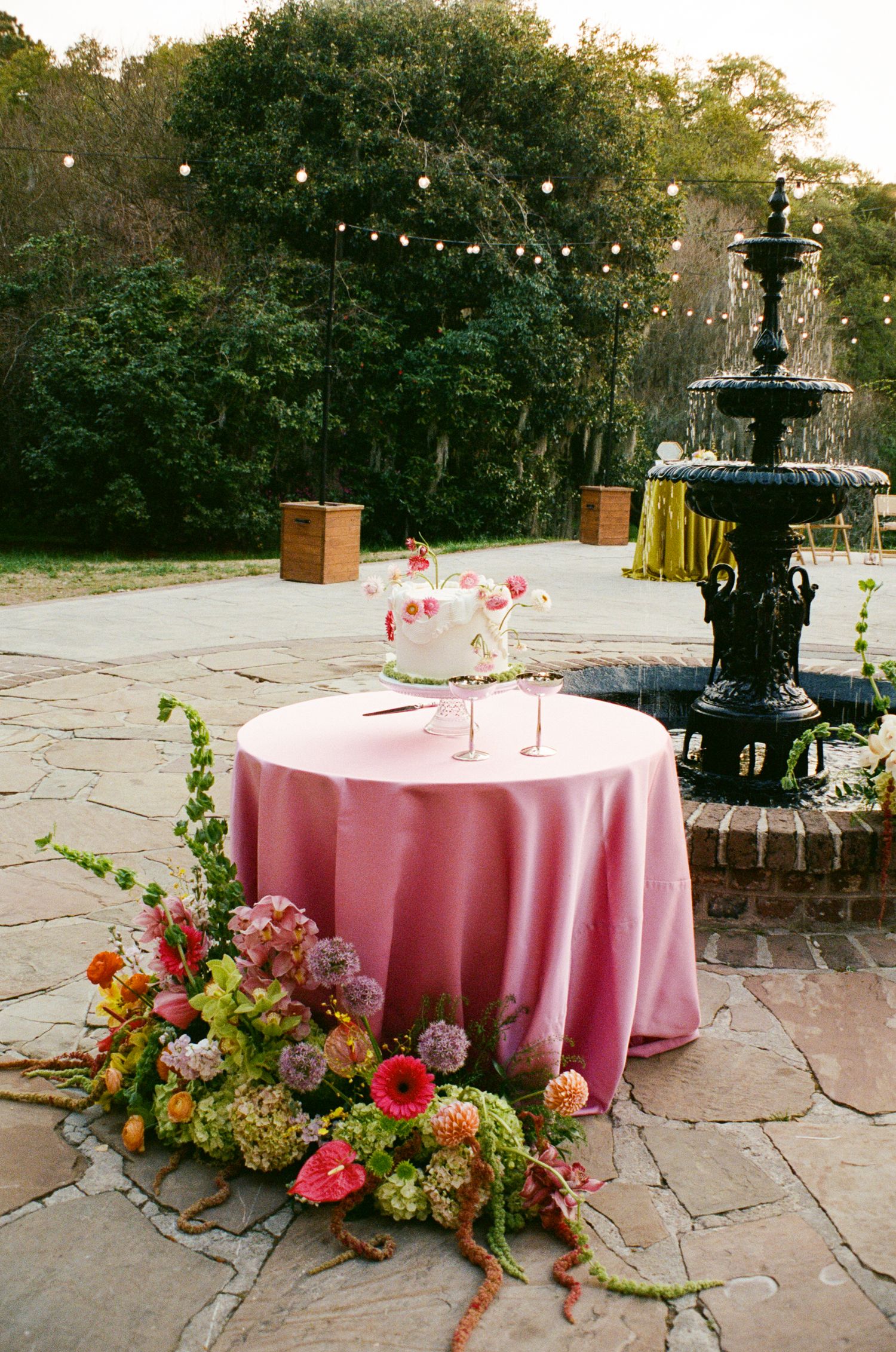 Outdoor wedding cake table with pink tablecloth and floral arrangement beside a black fountain on a stone patio.