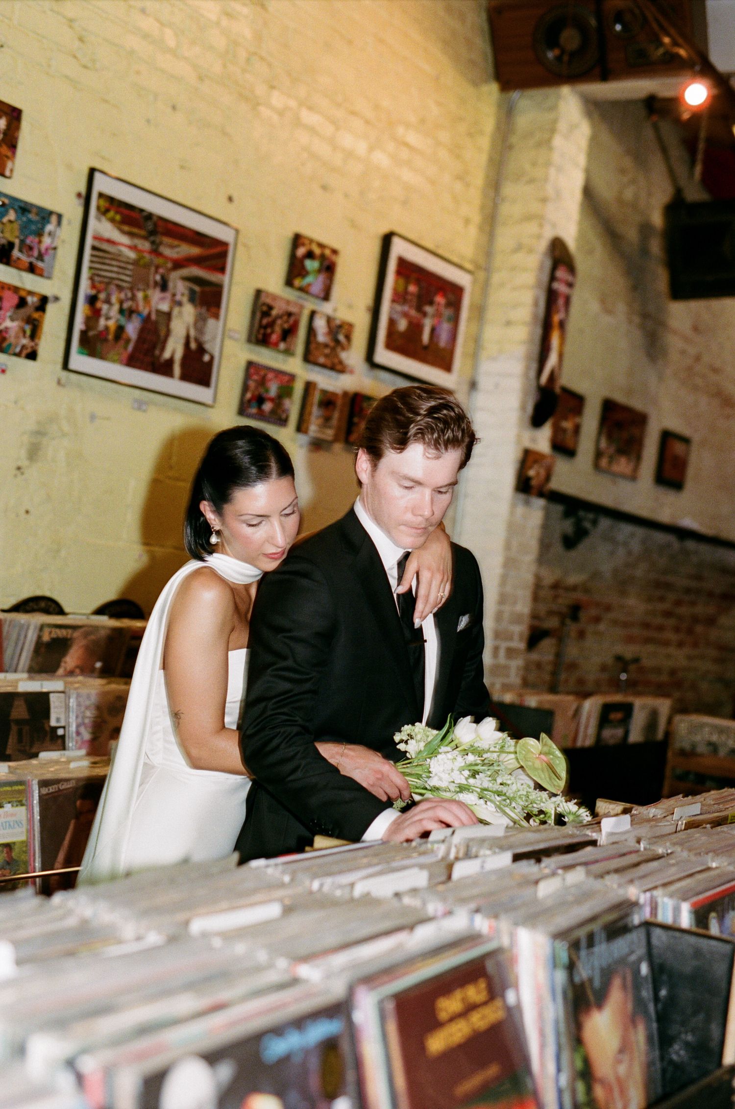 A bride in a white gown and a groom in a black suit with a bouquet browse vinyl records in a record store decorated with framed art.