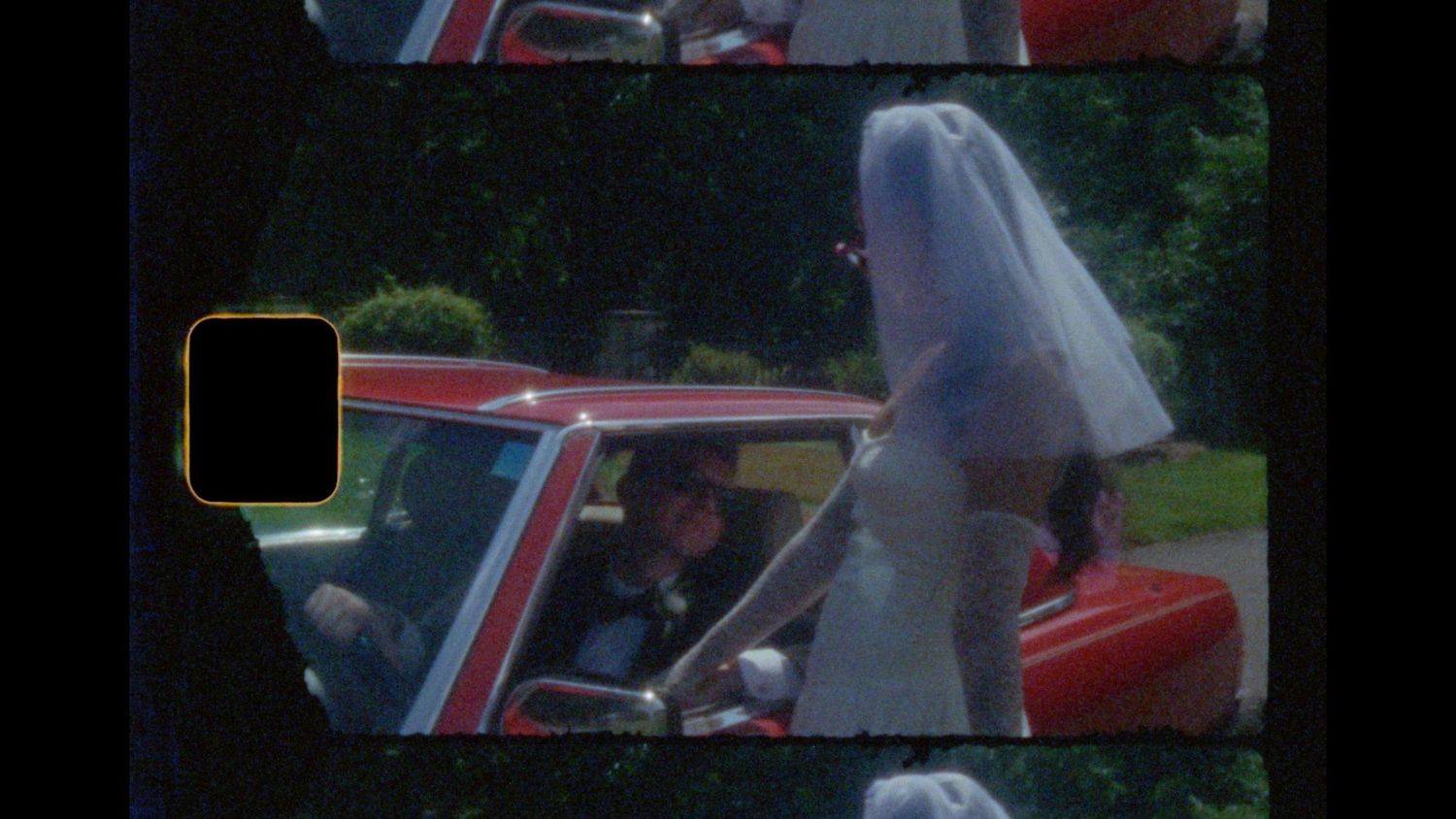 Bride in a white dress and veil leans toward a red classic car as a man smiles from the driver's seat, captured on vintage film.
