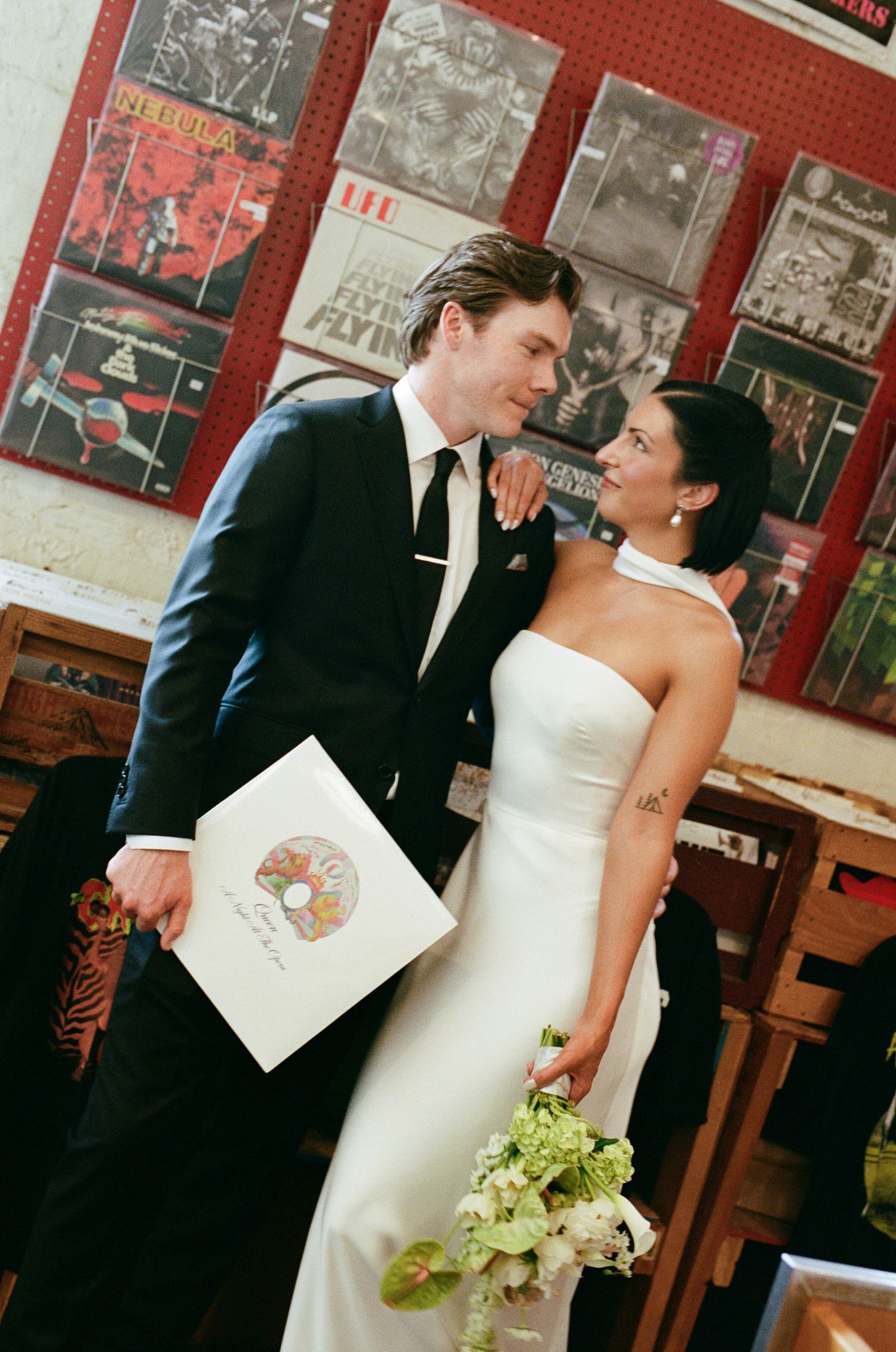 Bride in a strapless white gown and groom in a dark suit embrace beside a wall of vinyl records, holding a bouquet and a record sleeve.