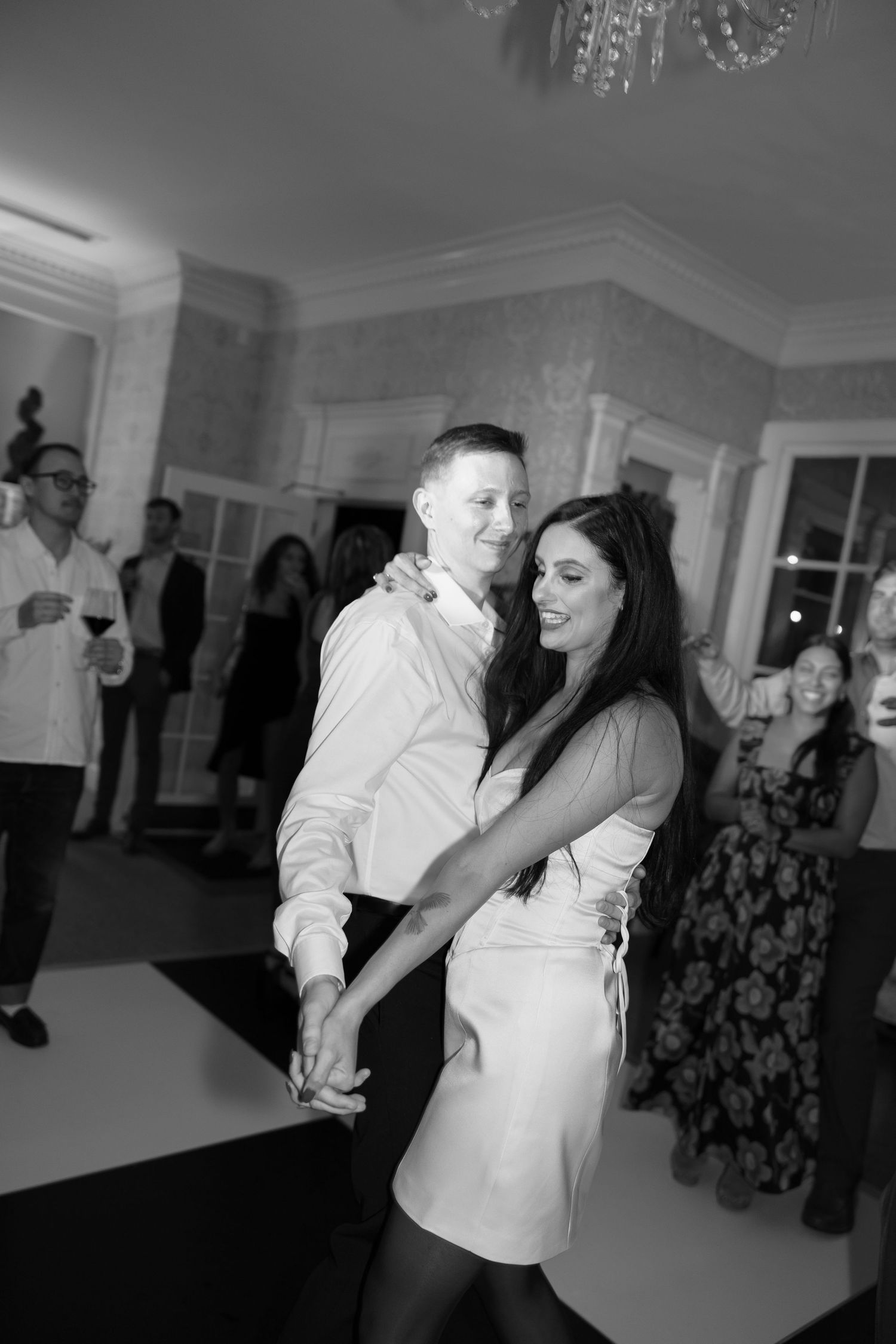 Black and white photo of a couple dancing and holding hands on a dance floor while guests watch at an elegant indoor wedding reception.
