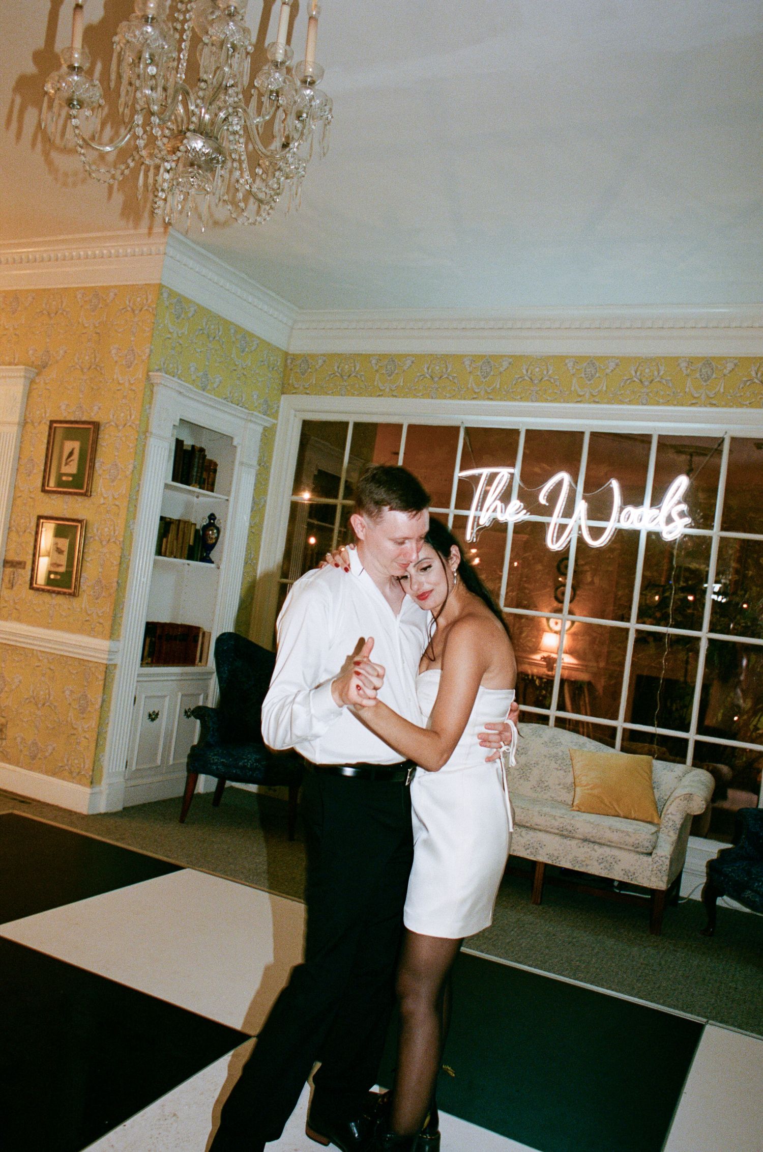 Couple in white dancing close on a black and white checkered floor in an elegant room with a chandelier and neon sign.