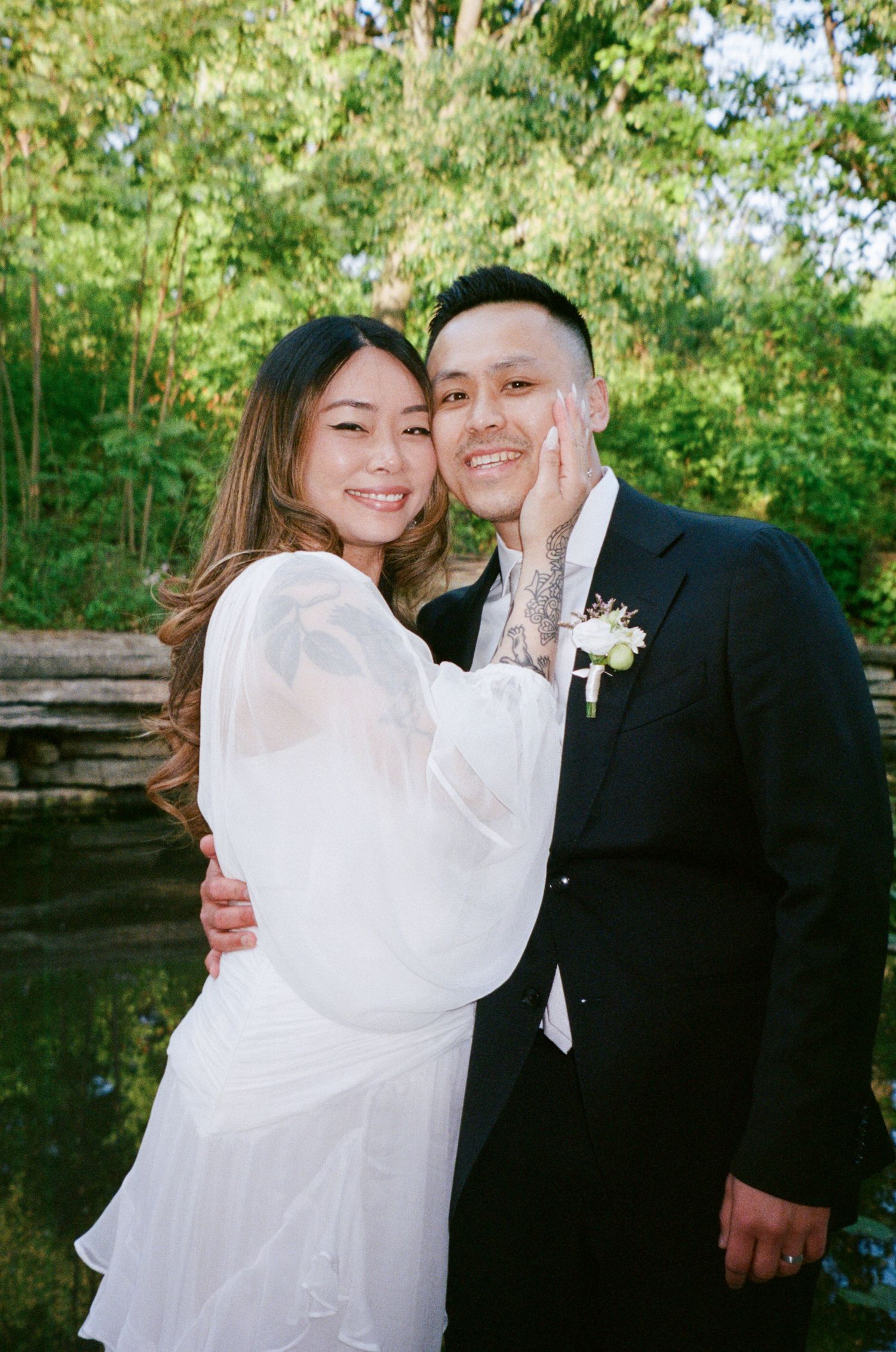 A couple in wedding attire embraces outdoors with trees and greenery in the background.