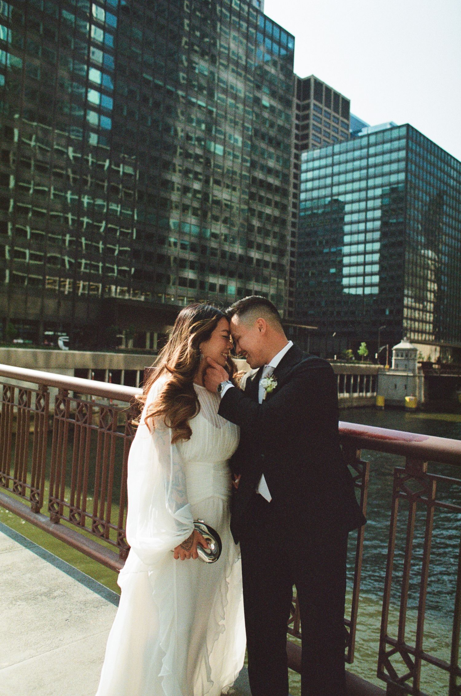 A romantic wedding couple embraces on a bridge with modern glass skyscrapers reflecting in the background.